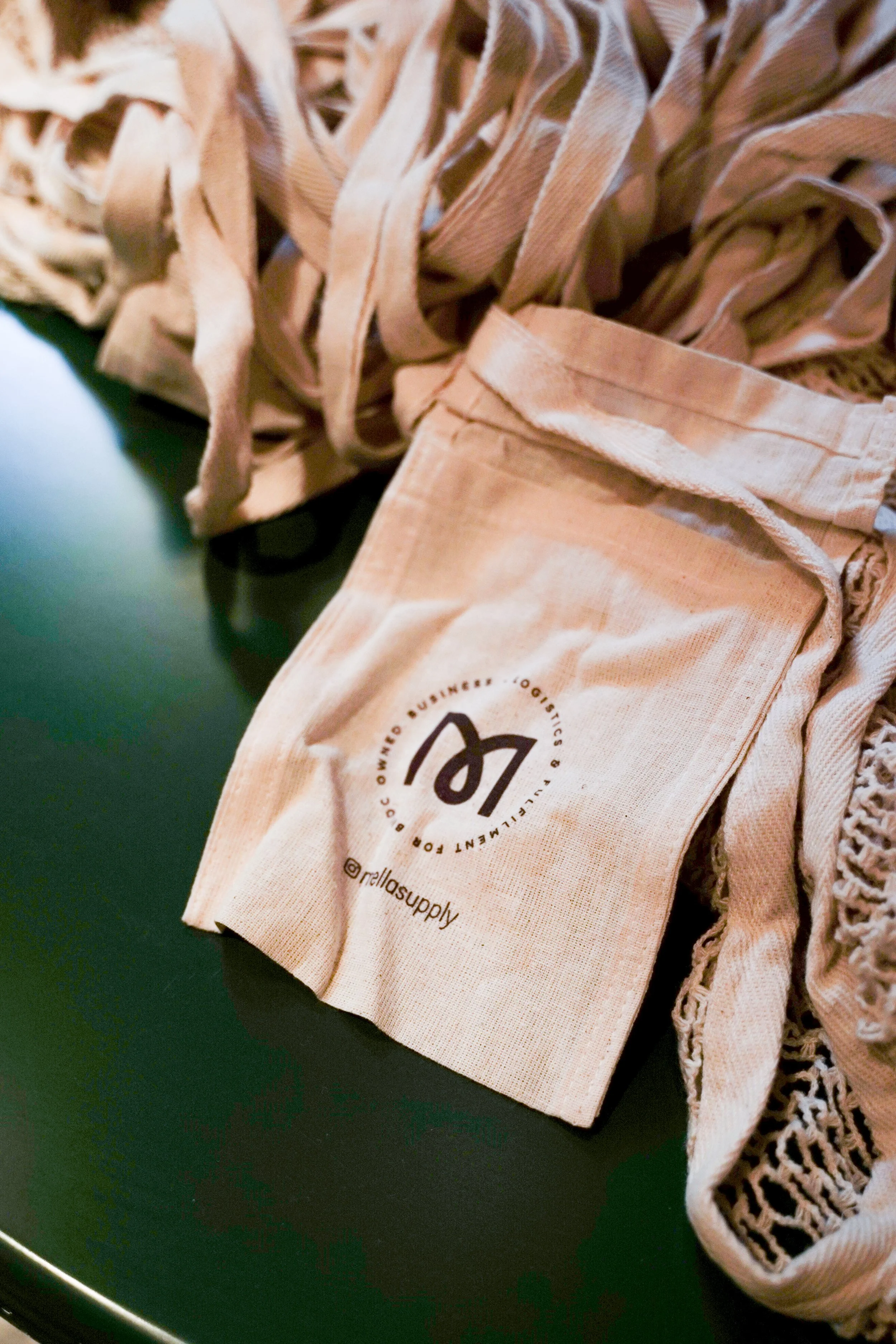 A beige fabric mask with a black logo and text that reads '@rellasupply' resting on a dark green surface, surrounded by beige fabric strips and lace.