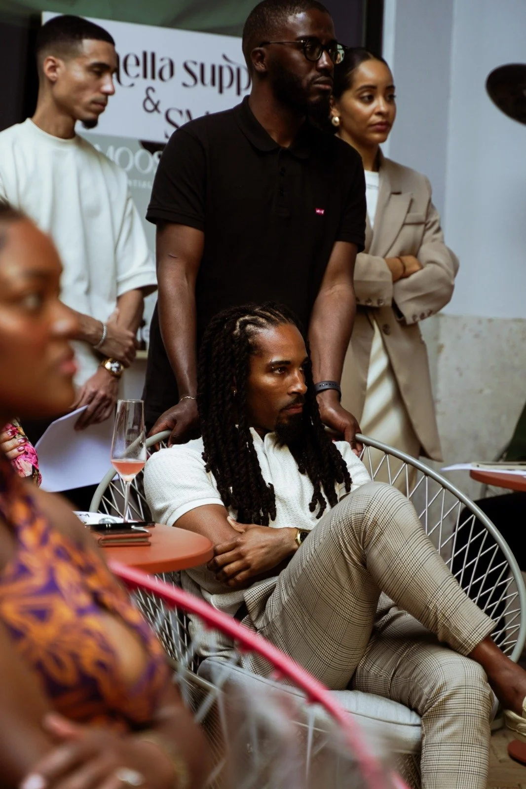 A group of people attending a meeting or event, with a man sitting in a chair in the foreground, and others standing behind, some taking notes. There is a sign in the background that partially reads "ella supp" and "S..."