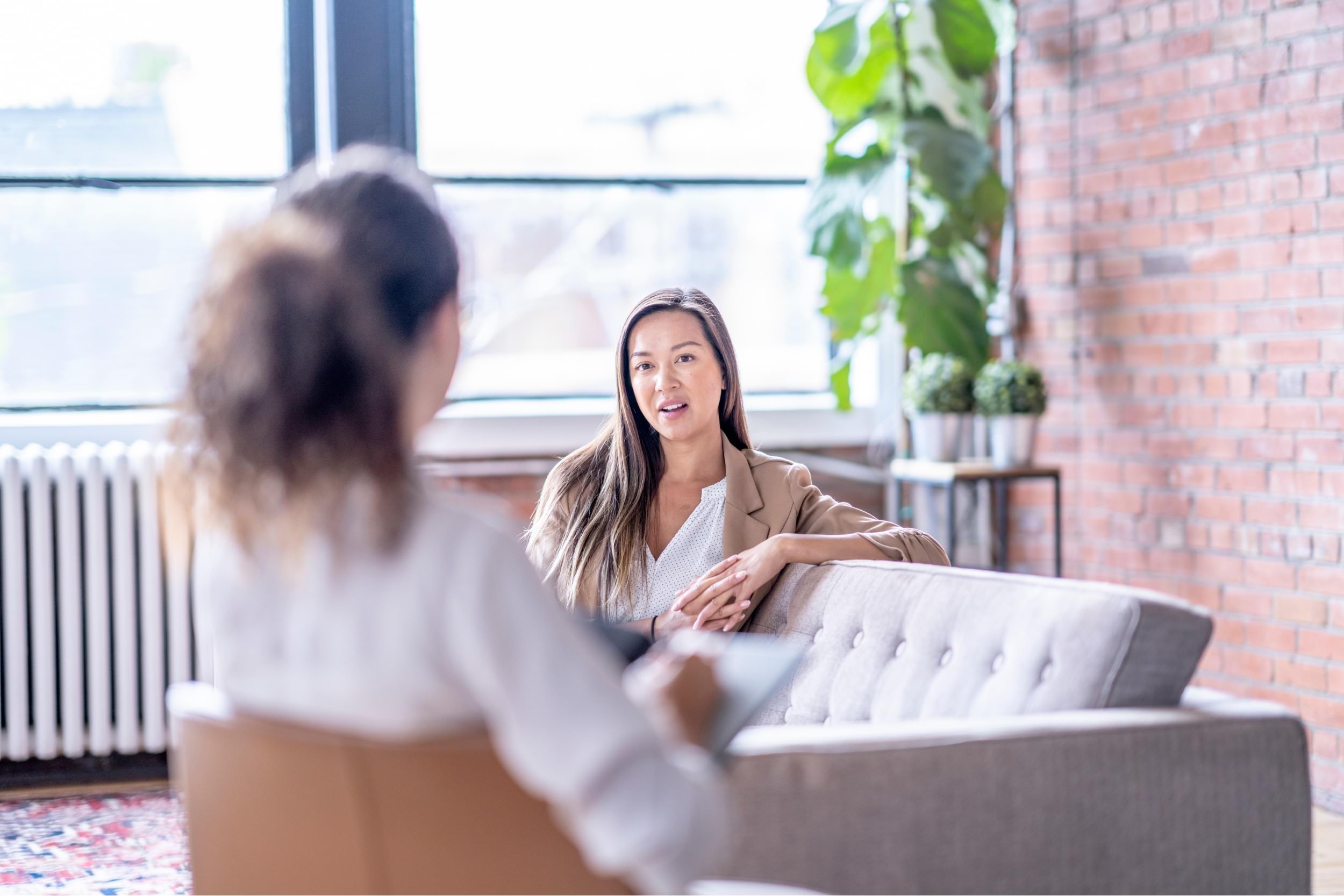 Two women having a conversation in a modern, cozy office with large windows, a brick wall, and indoor plants.