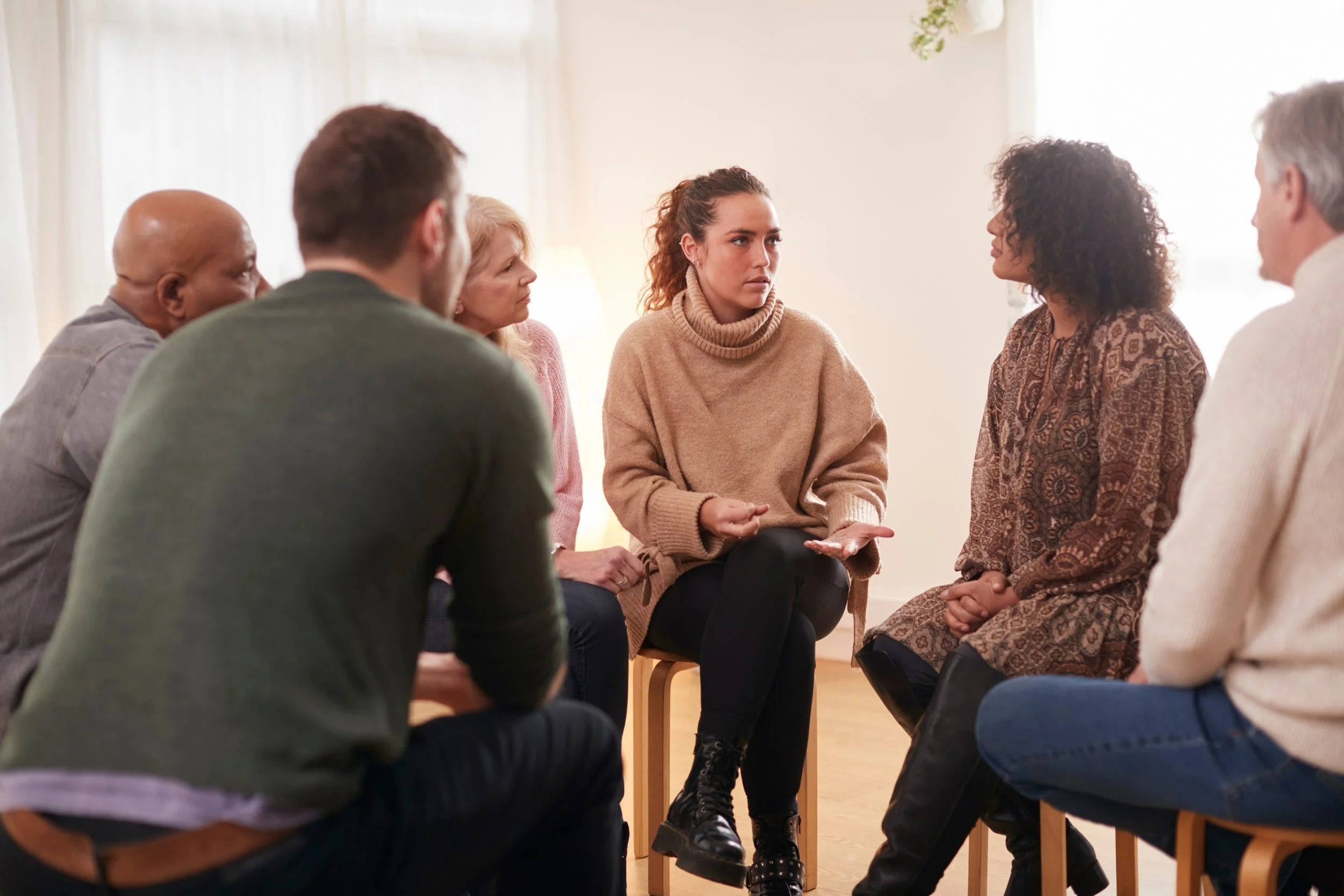 A diverse group of six people sitting in a circle in a cozy indoor space, engaged in a serious discussion with a young woman in the center speaking and others listening attentively.
