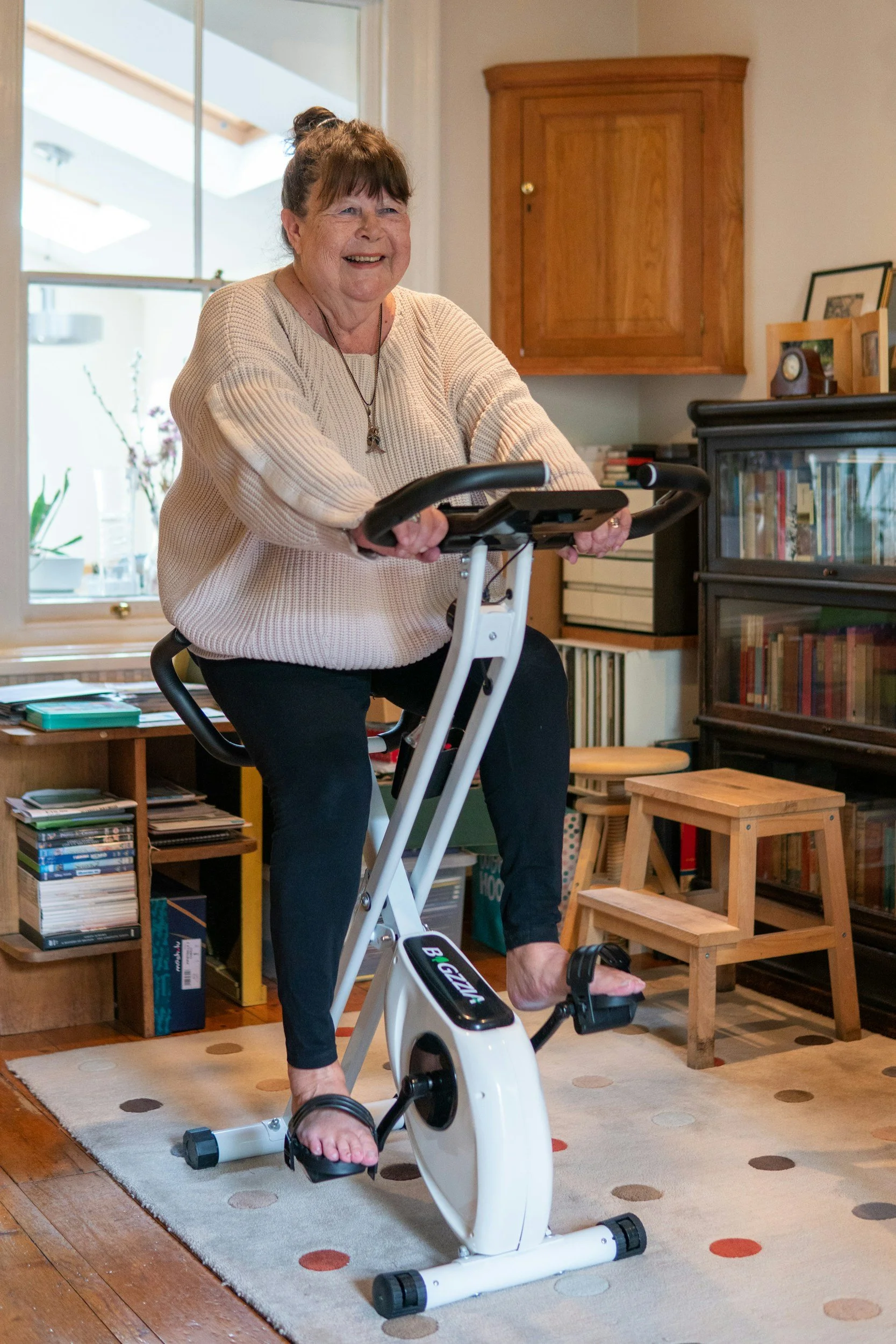 An elderly woman smiling while using a stationary exercise bike in a cozy living room.