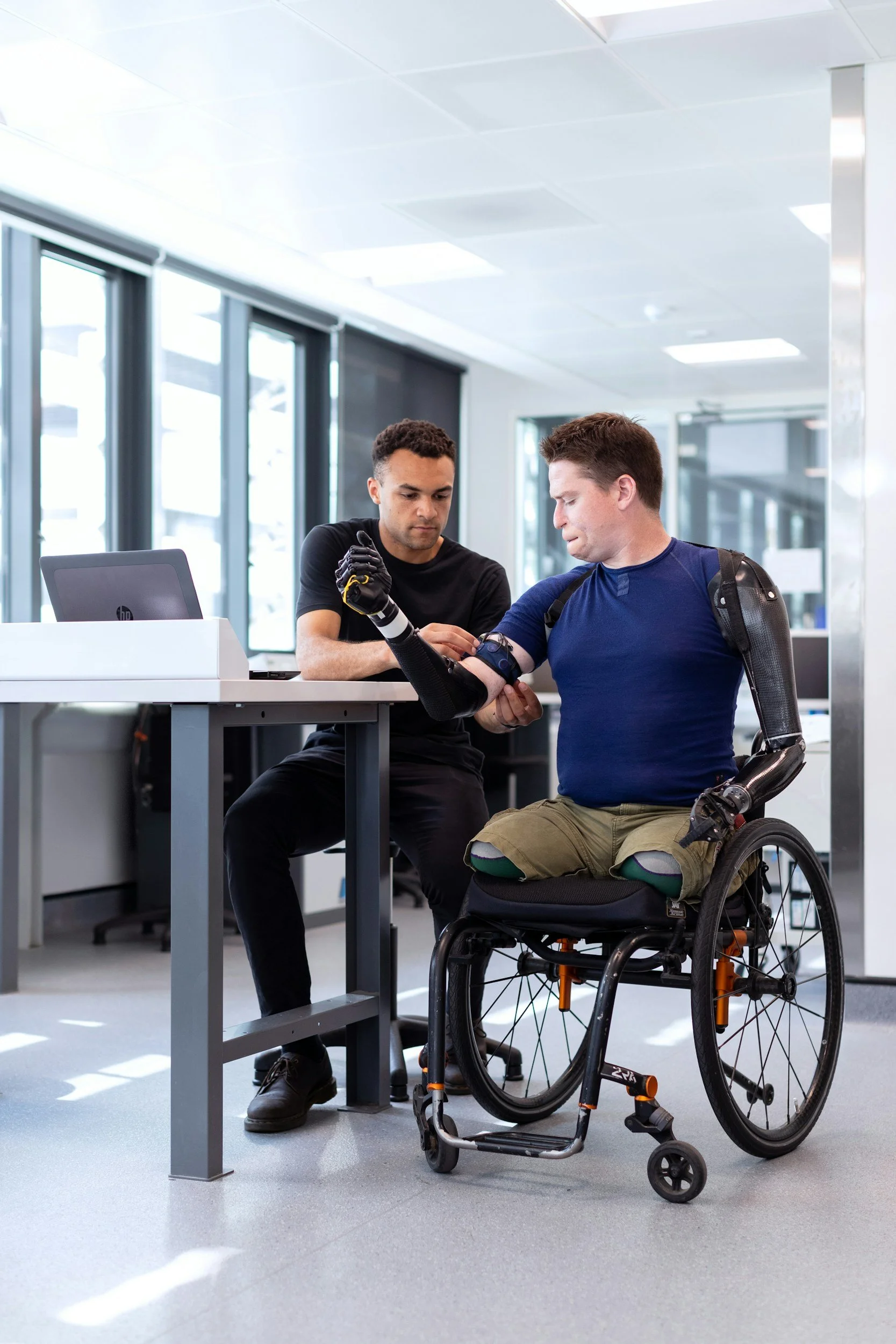 Two men, one with a prosthetic arm and the other with a wheelchair, are at a reception desk in a modern office, collaborating and looking at a device together.