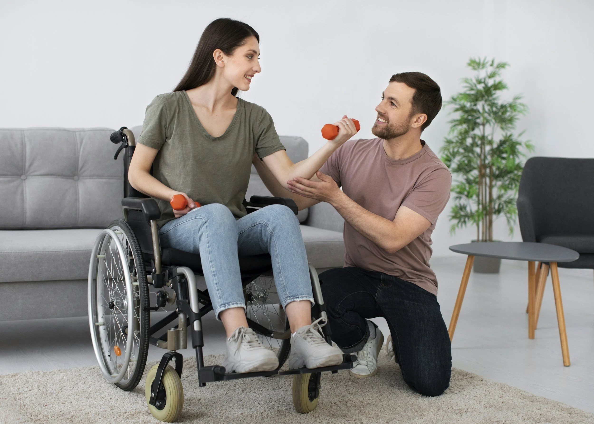 A woman in a wheelchair and a man are working out with small orange dumbbells in a living room. The woman is smiling and looking at the man, who is kneeling in front of her and holding her hand, both are smiling. There is a gray sofa and a potted plant in the background.