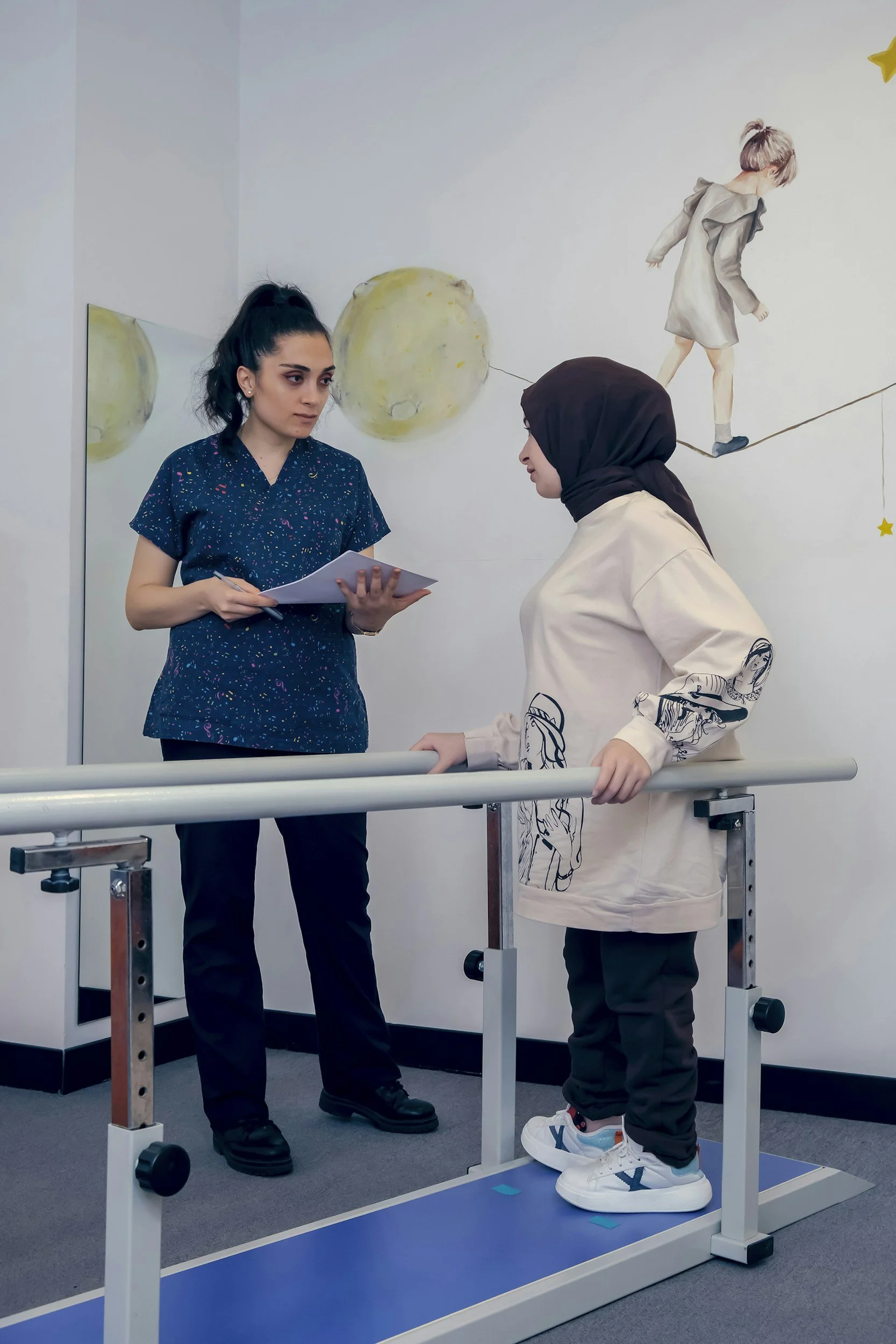 A young boy standing on a medical or rehabilitation balance board with support rails, engaging in balance exercises while a woman, possibly a therapist or nurse, discusses or instructs him. The scene takes place indoors, with a mural of a girl walking on a tightrope on the wall behind them.