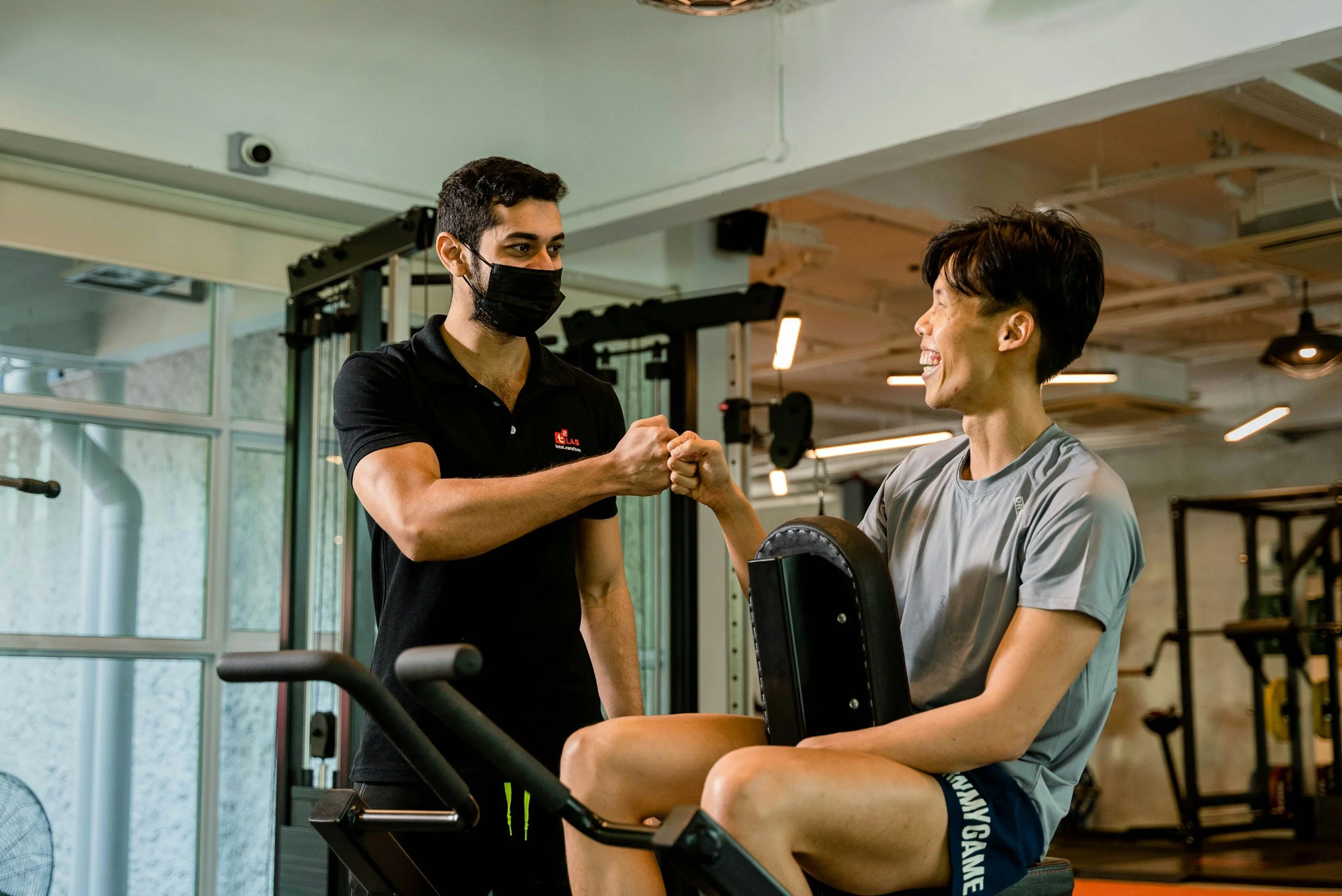A man wearing a black face mask and a black shirt with a logo, and a woman in workout attire, are greeting each other with a fist bump at a gym. The woman is seated on a workout machine, and they are smiling at each other.