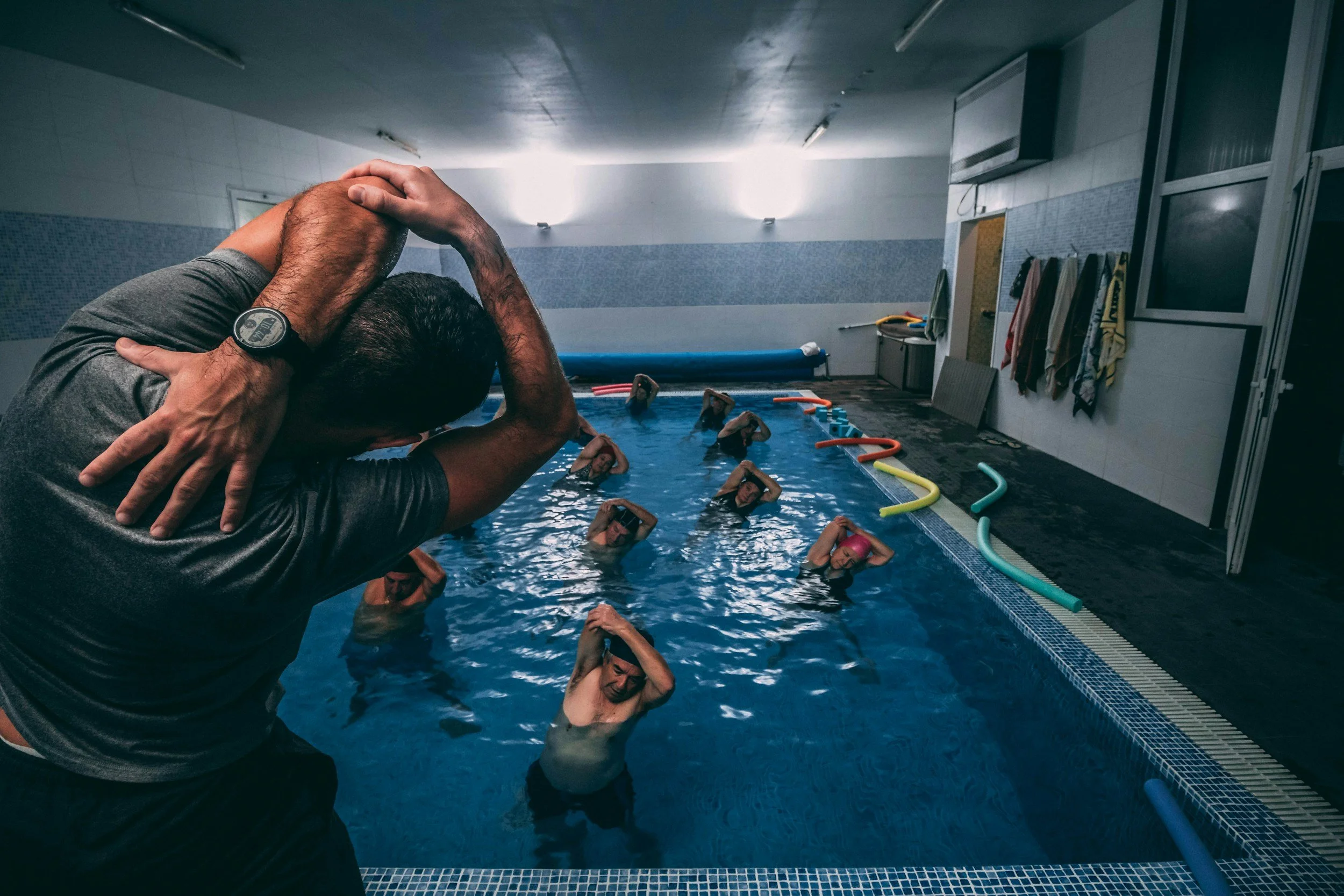 An indoor swimming pool with children participating in a swimming lesson. An instructor is in the foreground, holding and guiding a child's head in a swimming position. The children are practicing floating with their heads tilted back in the water. The pool area has tiled walls, pool noodles along the edge, and towels hanging on the wall.