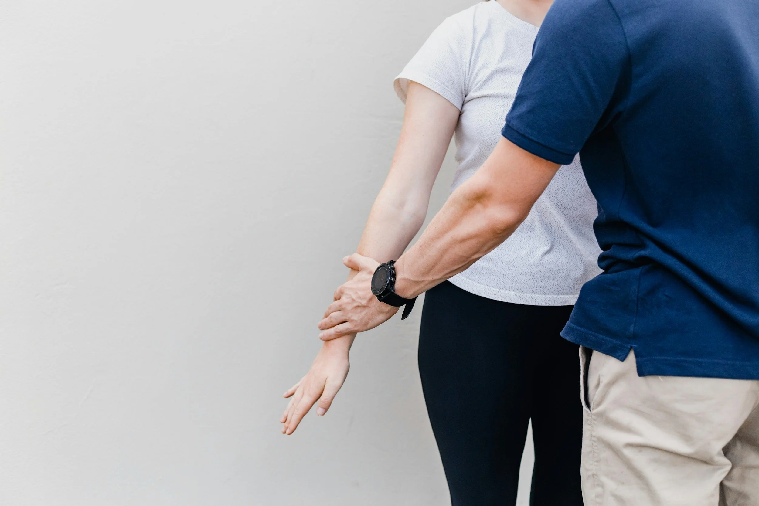 Two people, one in a white shirt and the other in a blue shirt, are holding each other's arms against a plain white wall.