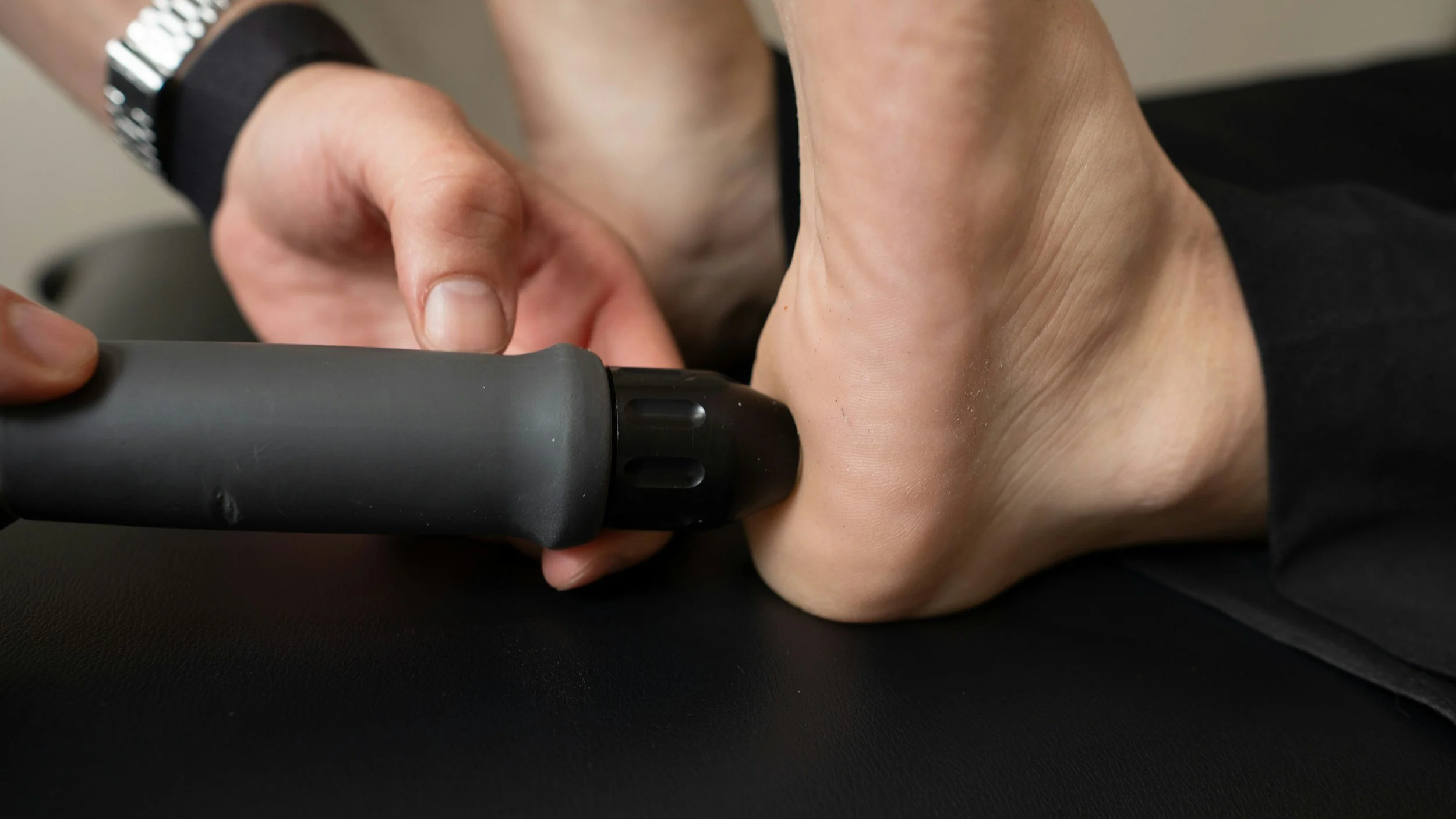 Close-up of a person using a massage gun on their foot, noting the person's hand, foot, and part of their wrist and watch.