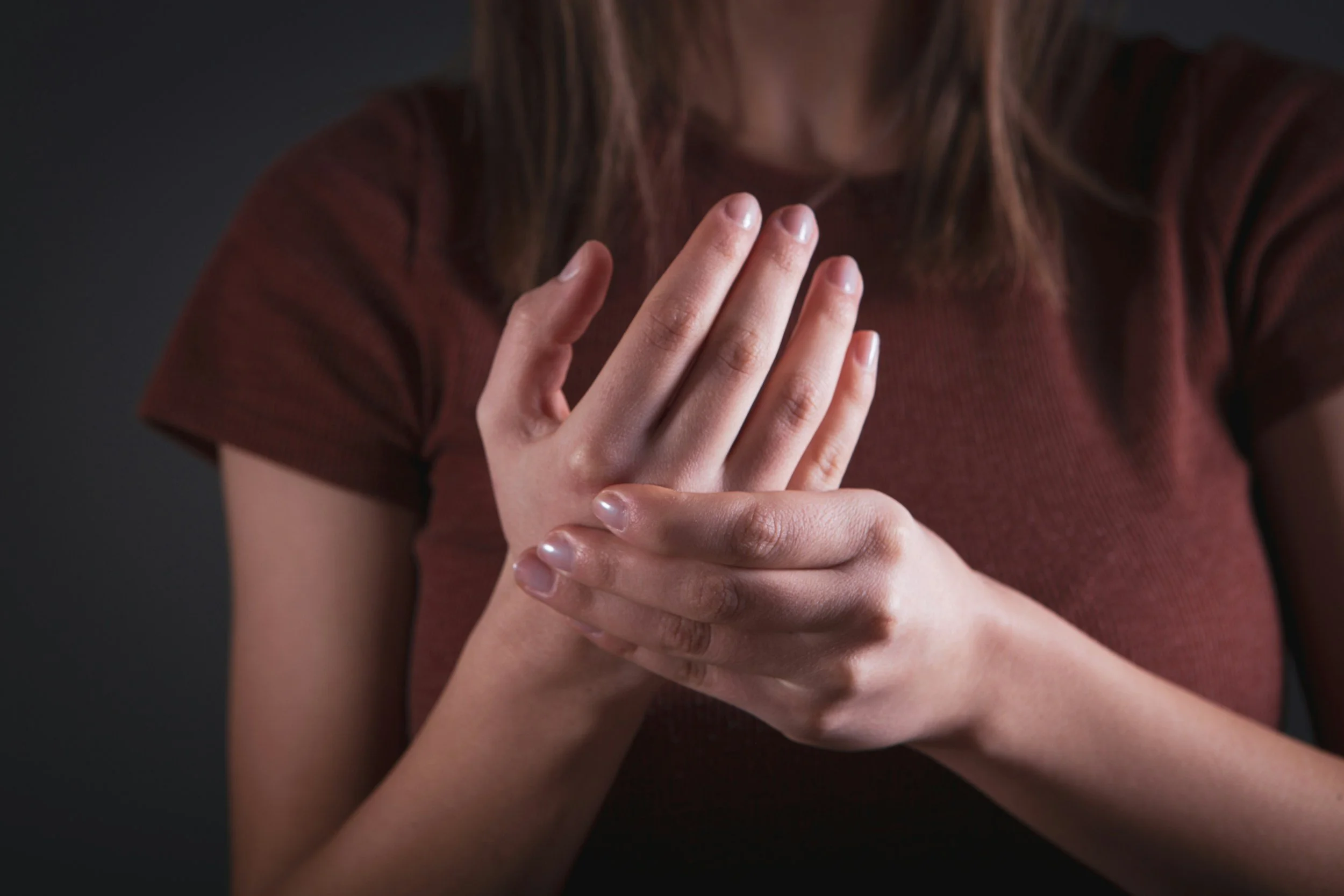 A person with long hair wearing a brown shirt is holding their hand up with fingers touching, against a dark background.