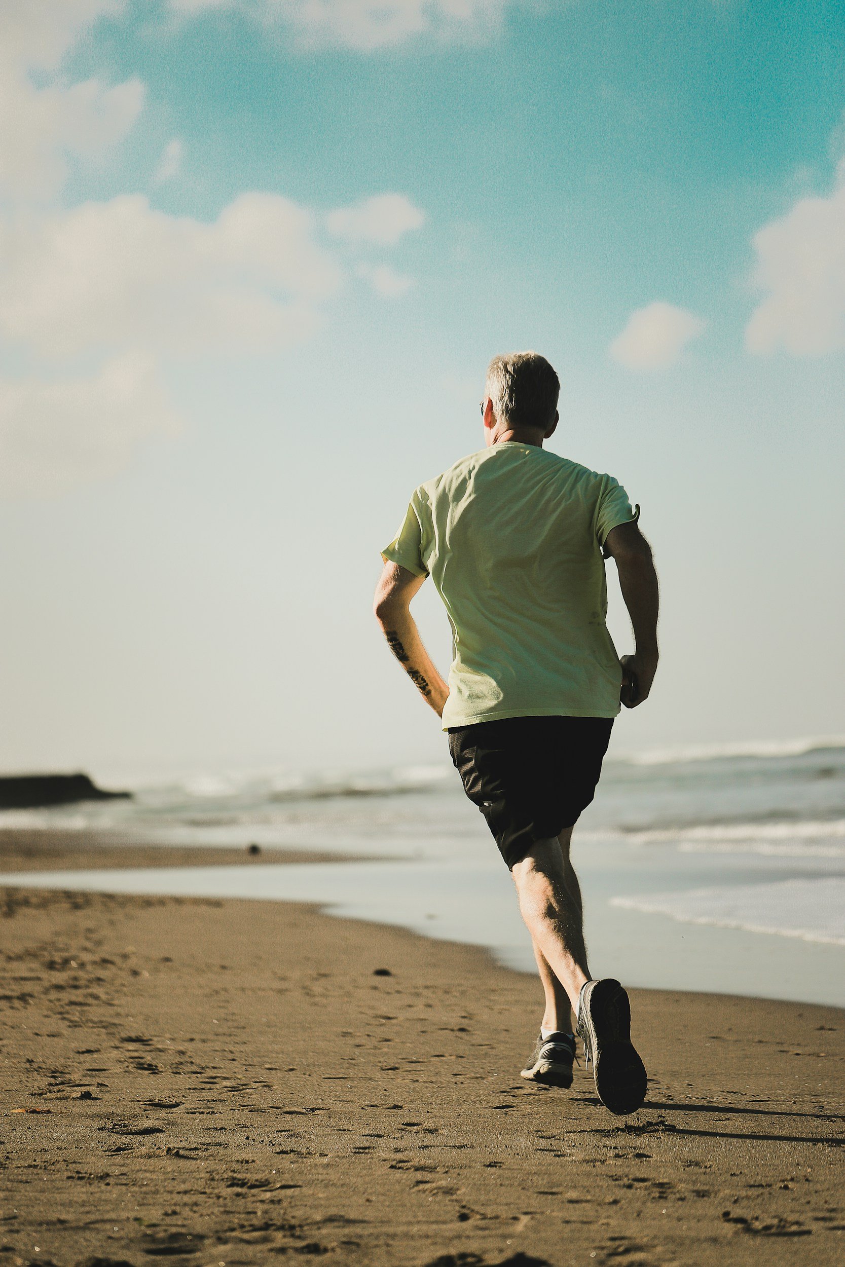 A man jogging on a sandy beach during the day, facing away from the camera, with clouds in the sky.