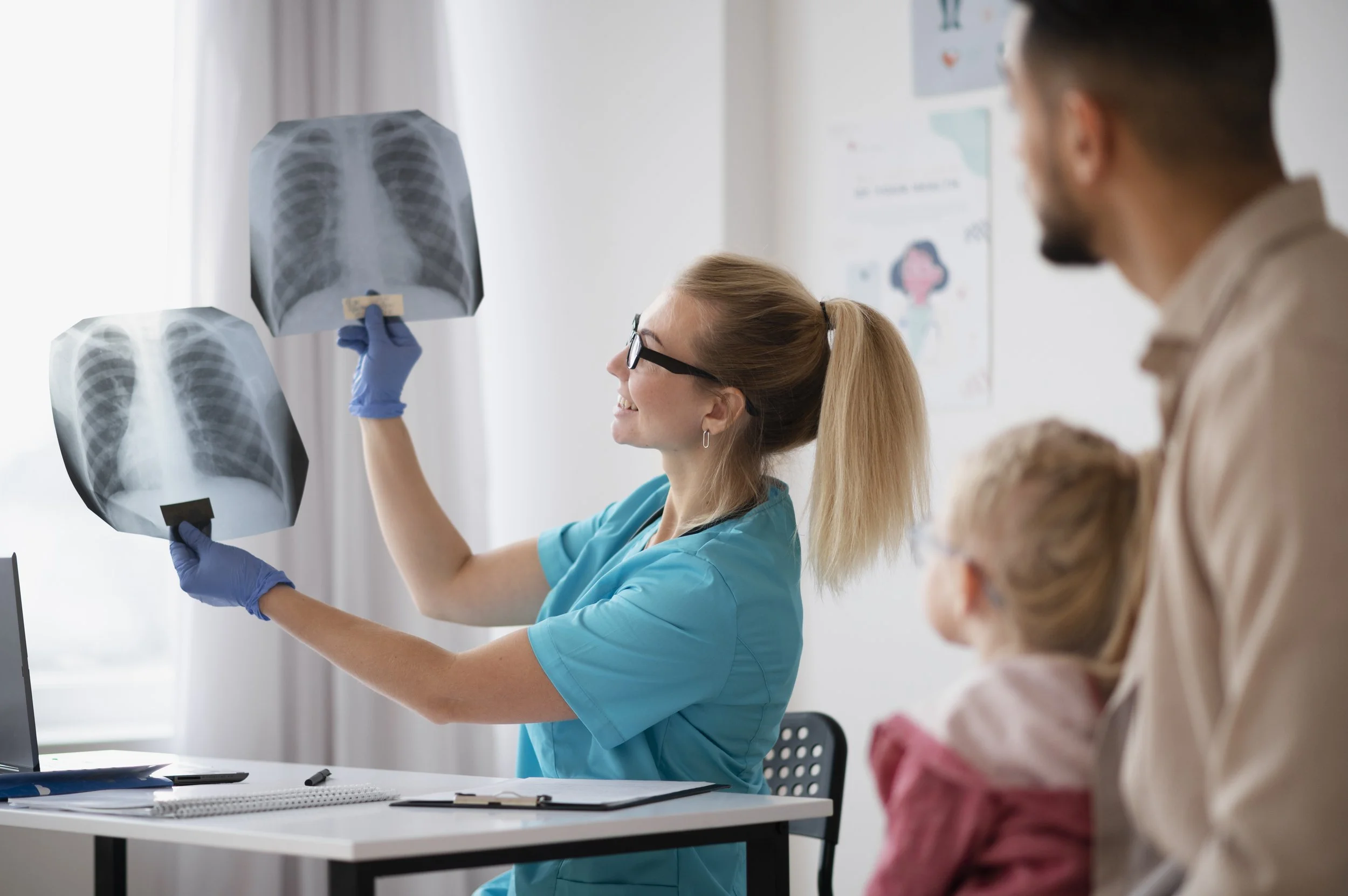 A healthcare professional in blue scrubs and gloves examining chest X-ray images held by another person in a medical office with people observing.