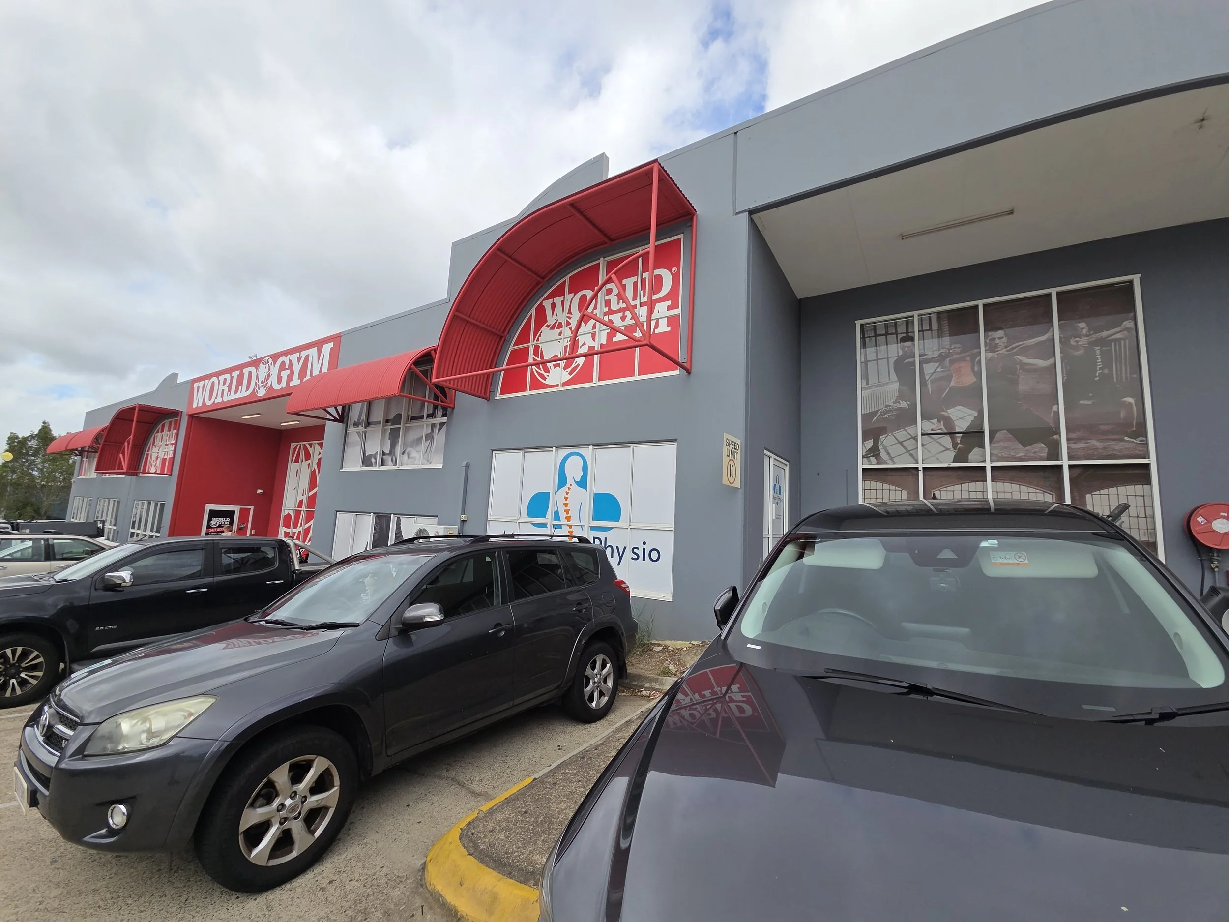 Exterior view of a gym with the sign 'World Gym,' featuring red awnings and large windows showing gym equipment and people working out inside. Several cars are parked in the parking lot.