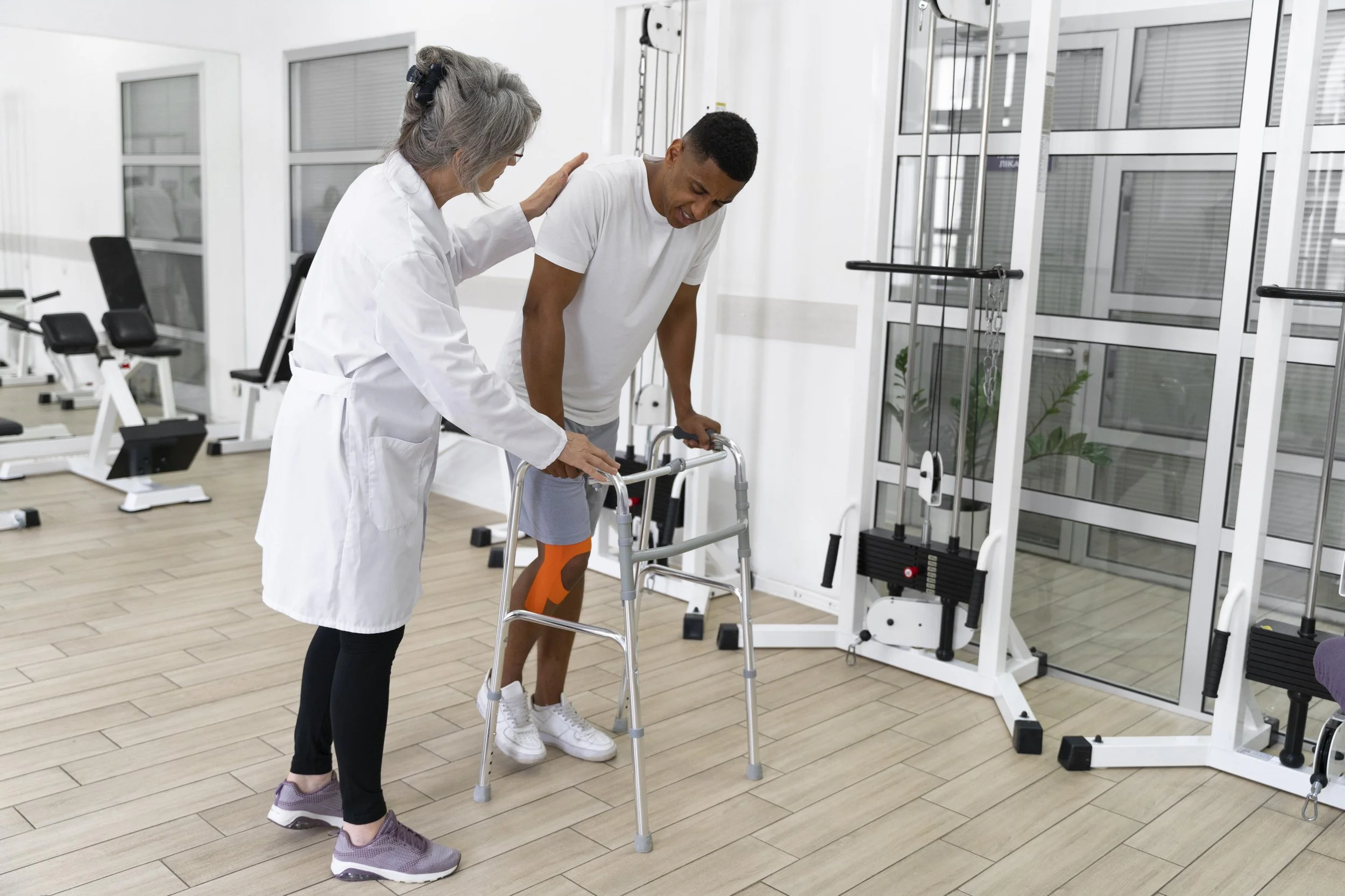 A healthcare professional helping a young man with leg injury to walk using a walker in a physiotherapy clinic.