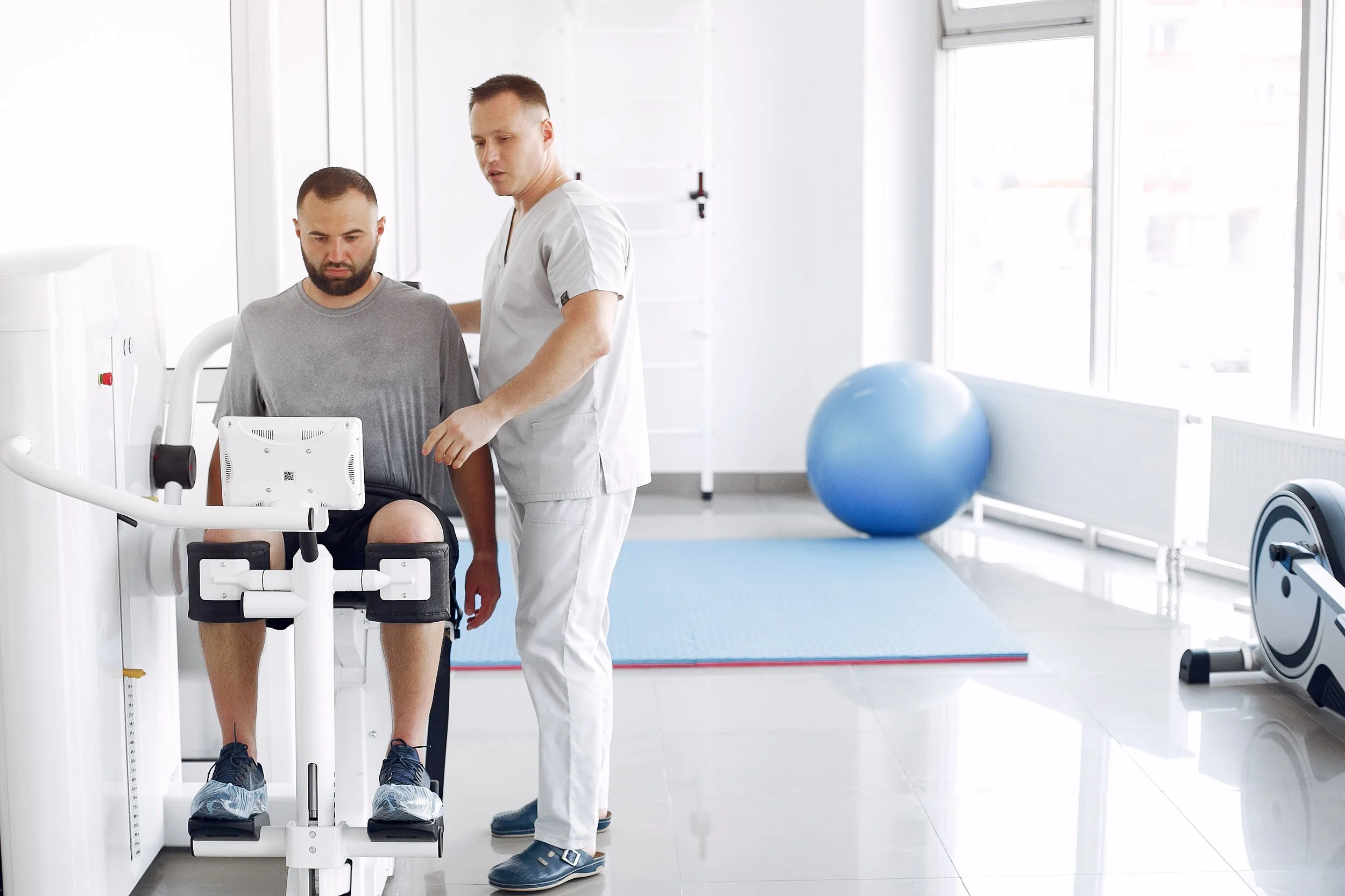 A man is seated on a medical exercise device in a bright, modern physical therapy or rehabilitation room. A healthcare professional in white scrubs is standing next to him, providing assistance. The room has large windows, a blue exercise ball, a rowing machine, and other fitness equipment.