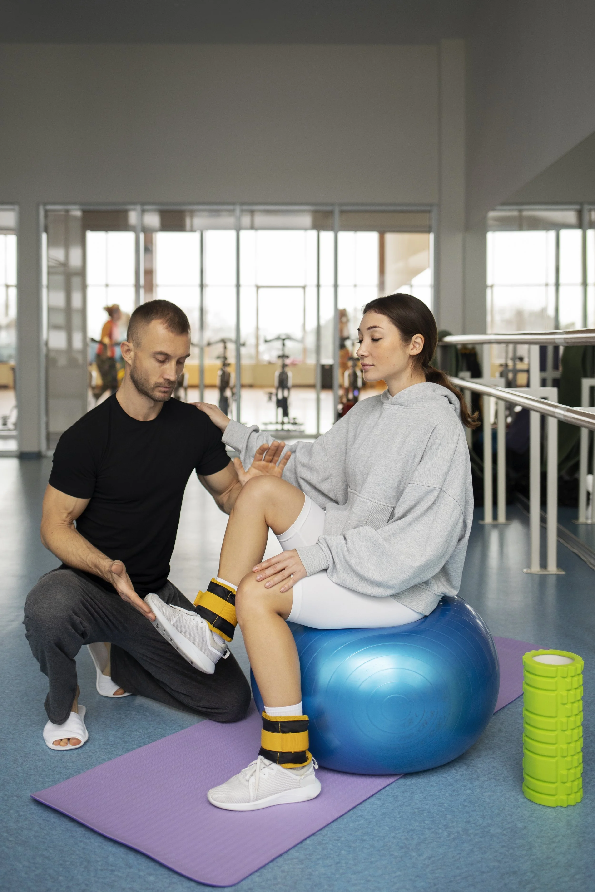 Physical therapist assisting a young woman with knee training on a stability ball at a gym.