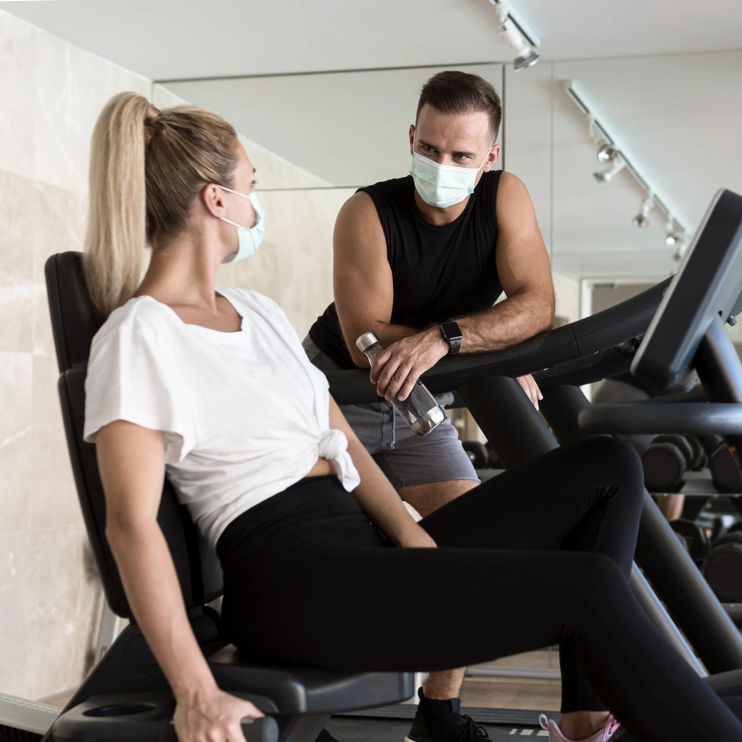 A man and a woman wearing masks at a gym or fitness center. The man is standing, leaning on a treadmill, and the woman is sitting on the treadmill.