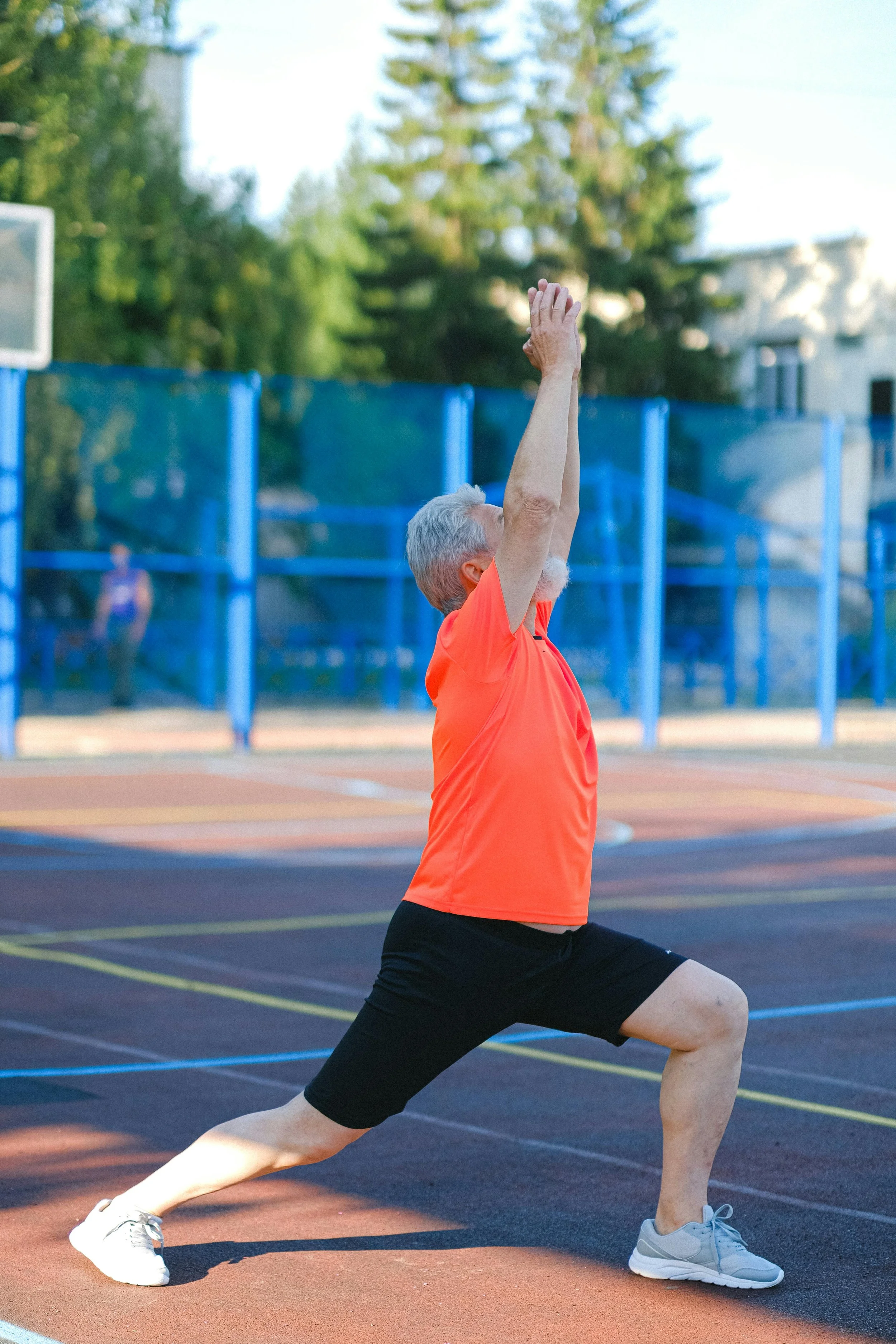 An older man with gray hair and beard doing yoga outdoors on a track, wearing an orange shirt and black shorts, in a lunge pose with arms raised, with a blue fence and trees in the background.