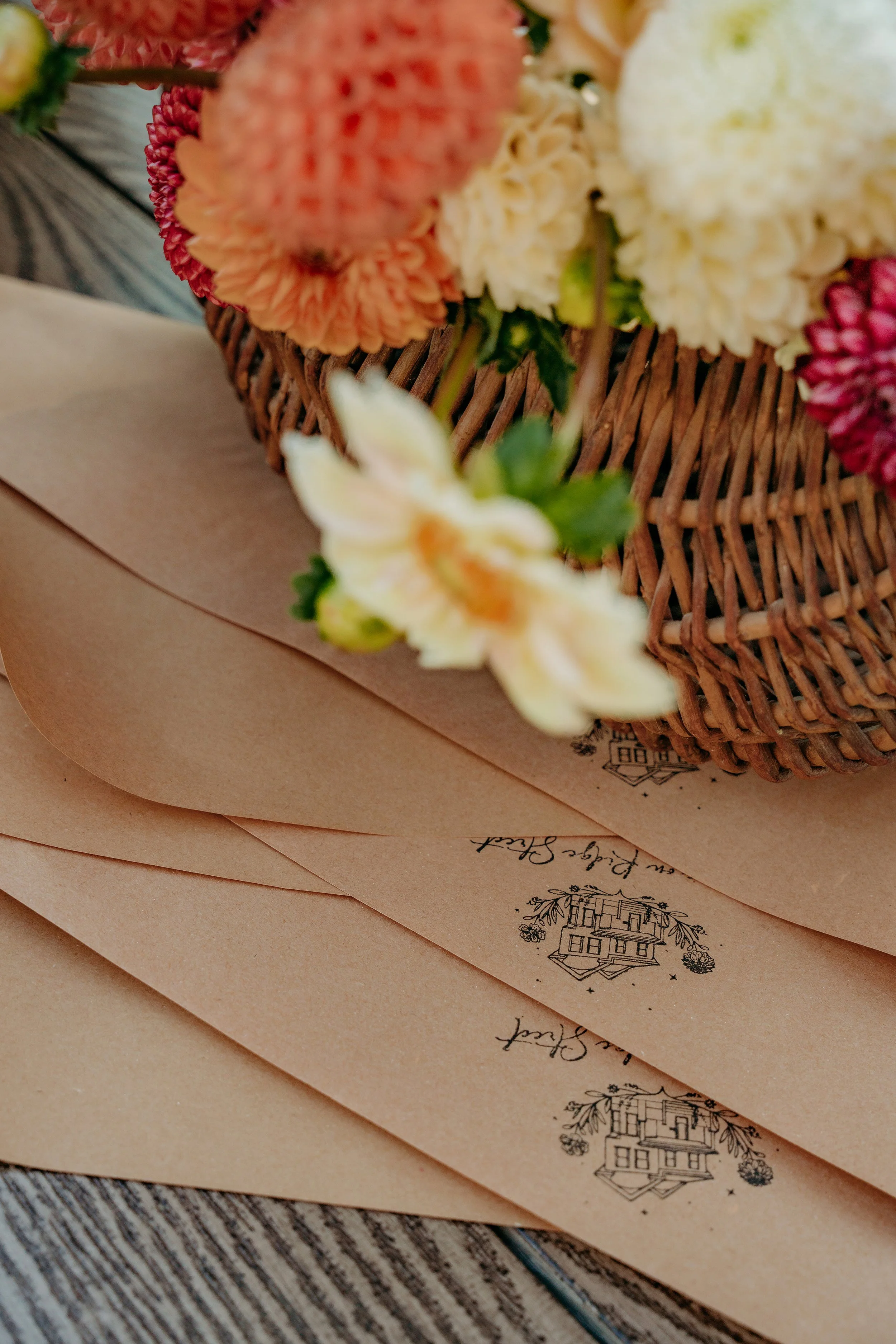 A wicker basket filled with flowers, including white, peach, and pink blooms, resting on tan paper envelopes with printed designs, on a wooden surface.
