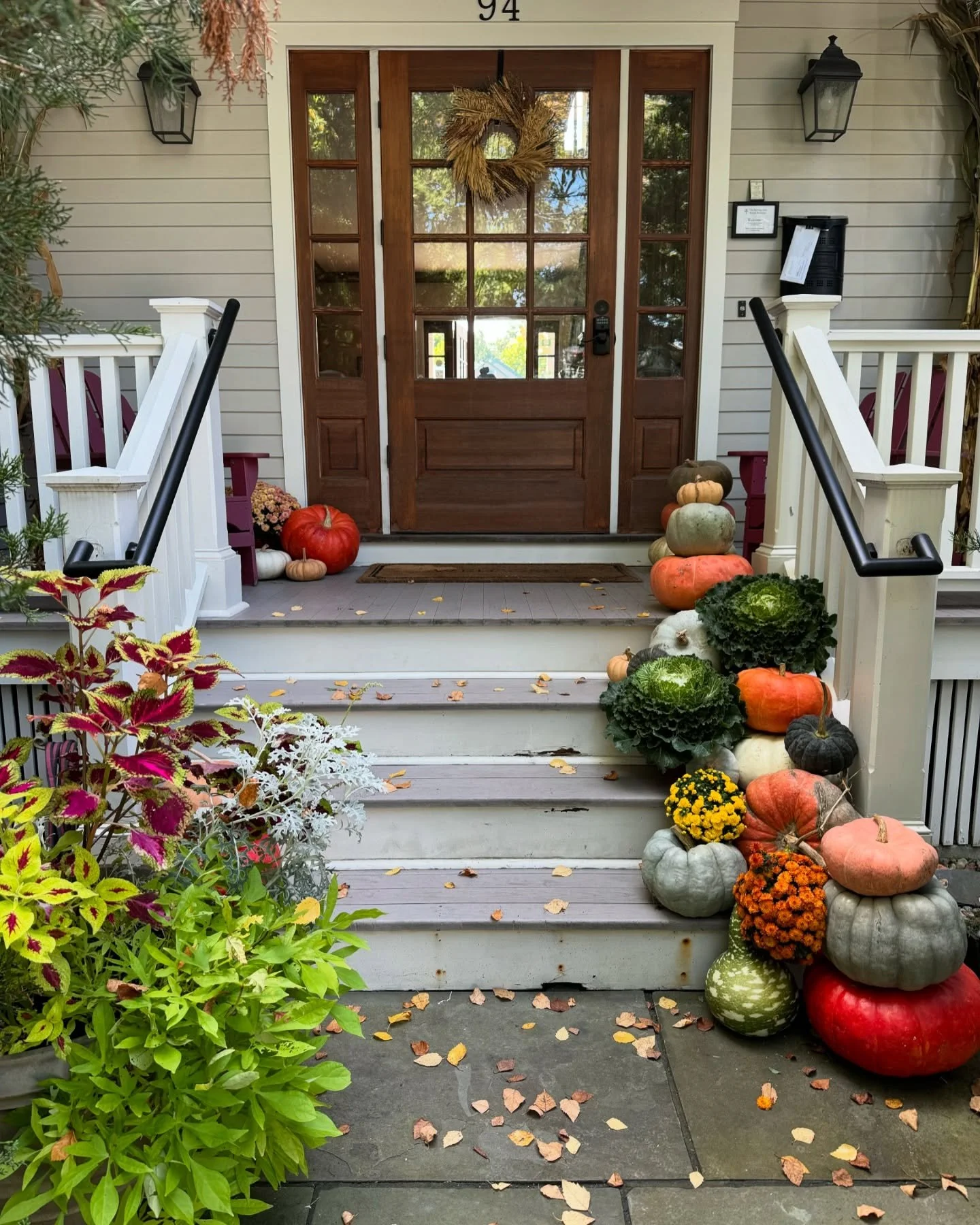 Front porch with a wooden door decorated with a wreath and surrounded by fall decorations including pumpkins, gourds, and flowers on steps and sides, with scattered leaves on the ground.
