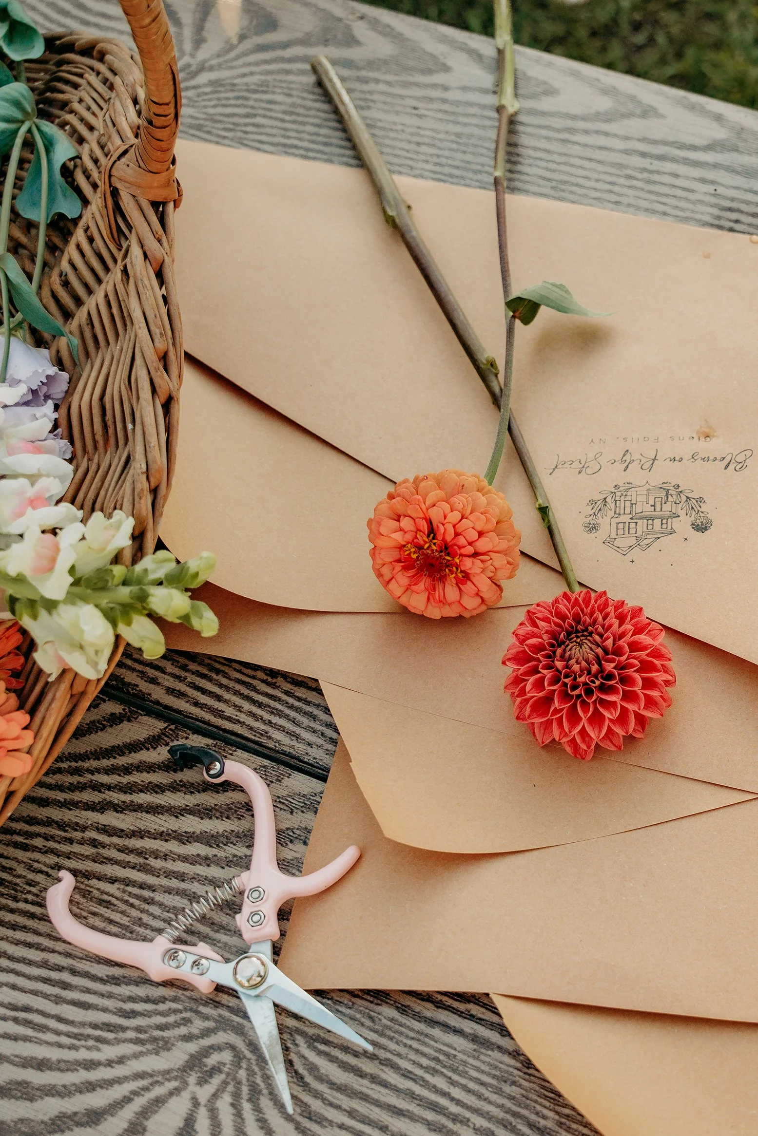Two colorful flowers resting on wedding invitations, next to a pair of pink scissors, with a wicker basket holding flowers in the background, all on a wooden surface.