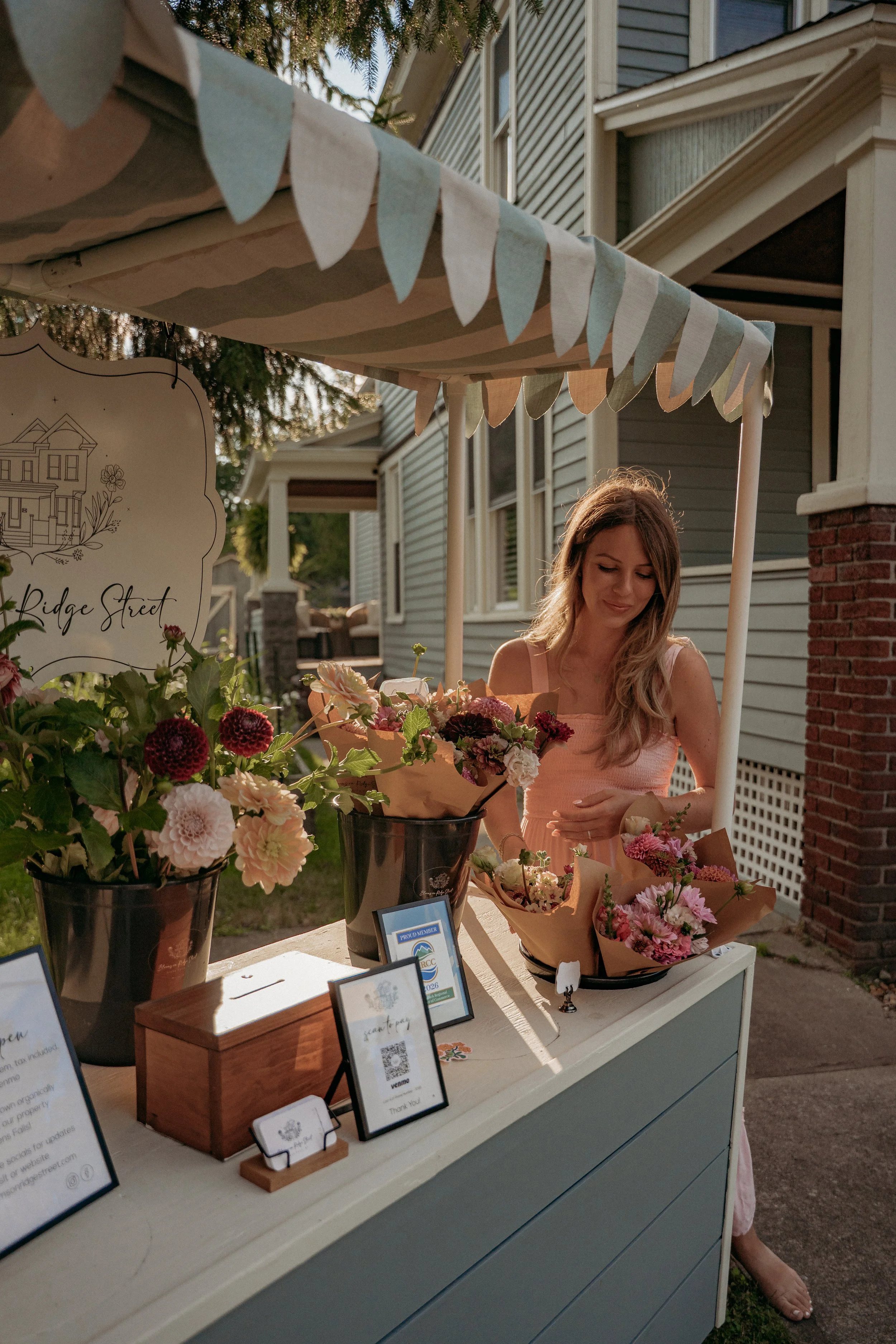 A bouquet of orange, peach, and white flowers in a black pot on a wooden table outdoors in front of a house with trees and a woman in the background.