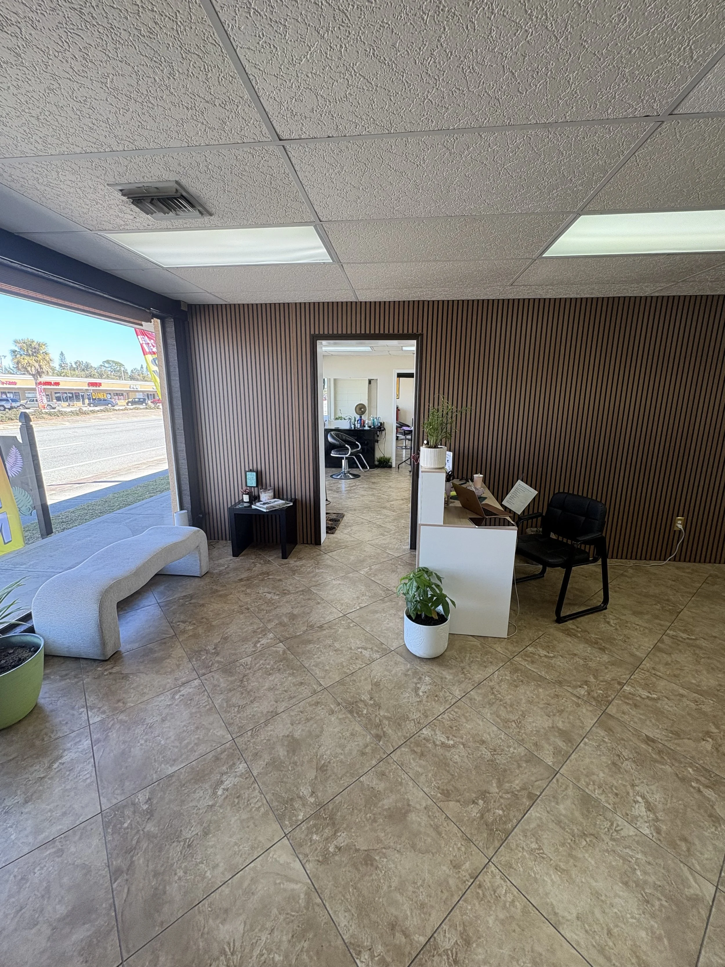 Clean, modern interior of a waiting or reception area with a white curved bench, a potted plant, and a reception desk. There is a doorway leading to a back room.