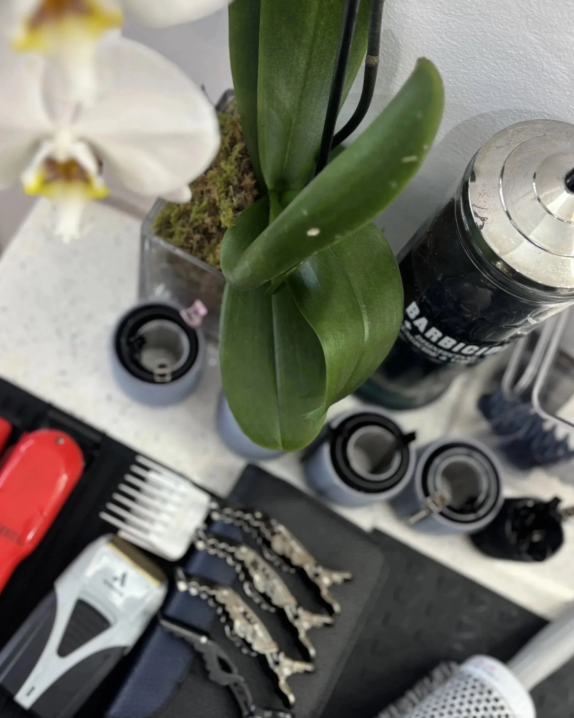 Top view of a workspace with a white orchid in a glass pot, a black container labeled 'Barbieri', a tall metal shaker, a black mat with hair clips, a red-handled tool, a hair clipper, and hair clips.