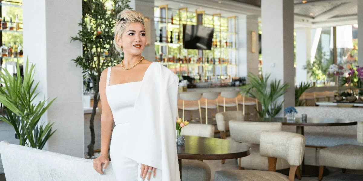 A woman with short blonde hair and jewelry in a white dress and jacket standing inside a modern restaurant or cafe, with tables, chairs, green plants, and a bar area in the background.