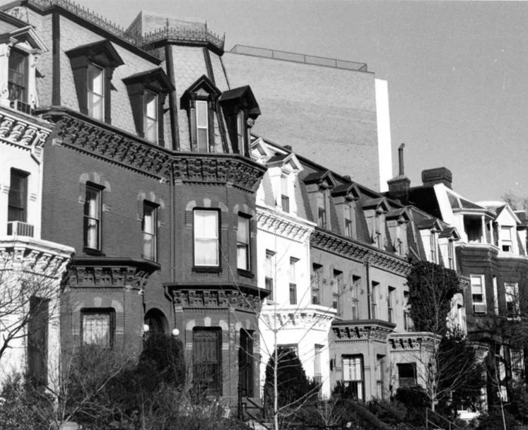 Black and white photo of row houses with distinct Victorian architectural features, including bay windows, ornate trim, and dormer windows on the upper floors.