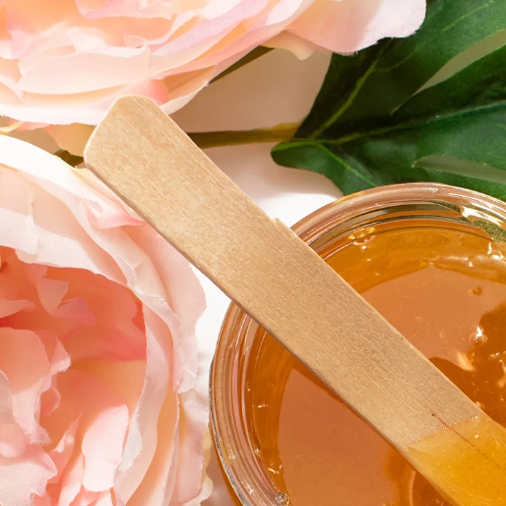 Glass jar of honey with a wooden honey dipper resting on top, pink peony flowers, and green leaves in the background.