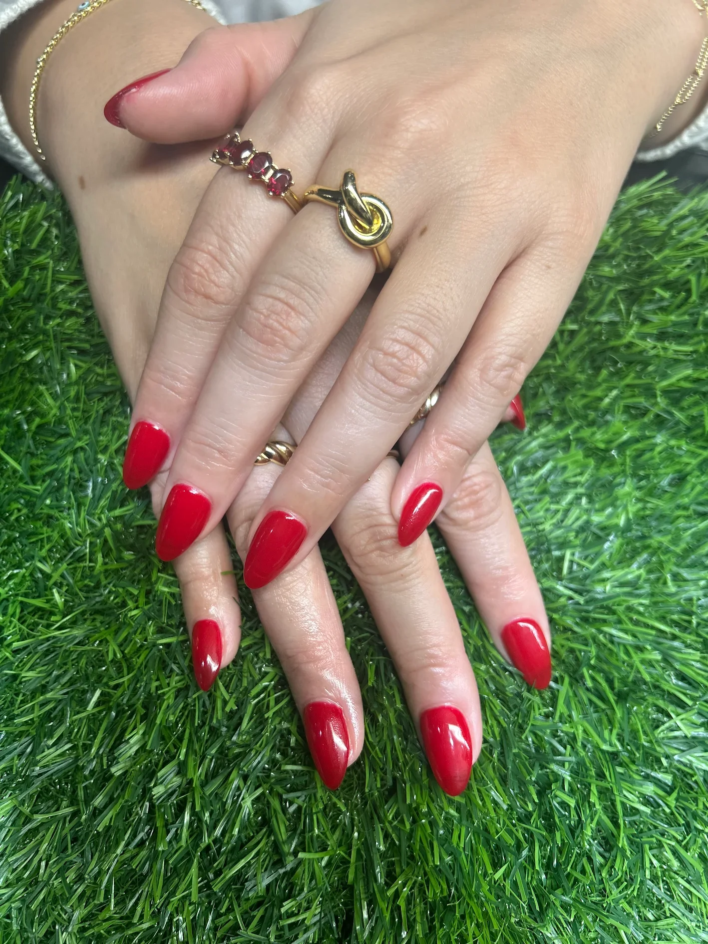 Hands with red painted nails, wearing jewelry, on green grass.