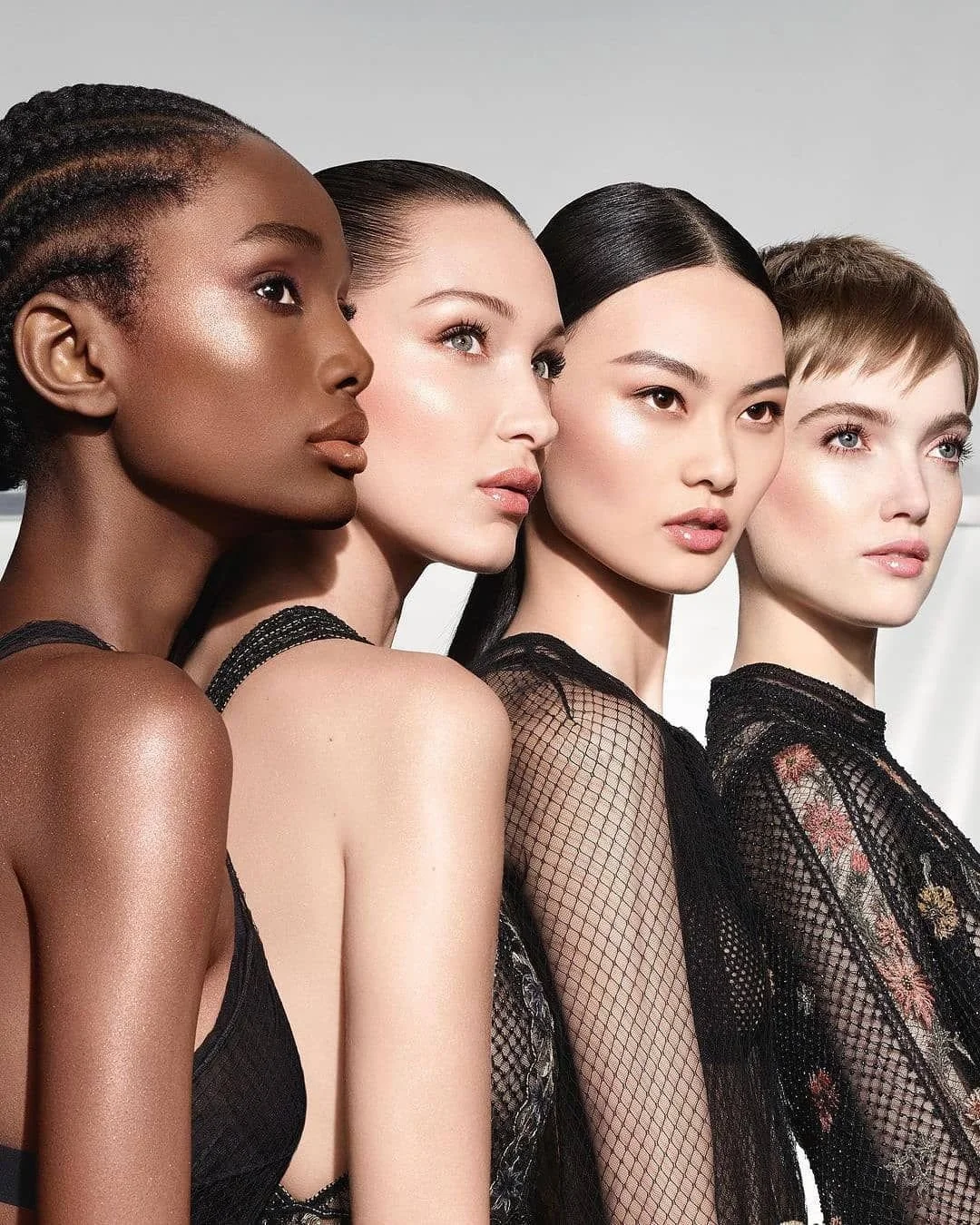 Four diverse women with different skin tones and hair styles wearing black lace and mesh clothing, posed closely together in a fashion photoshoot.