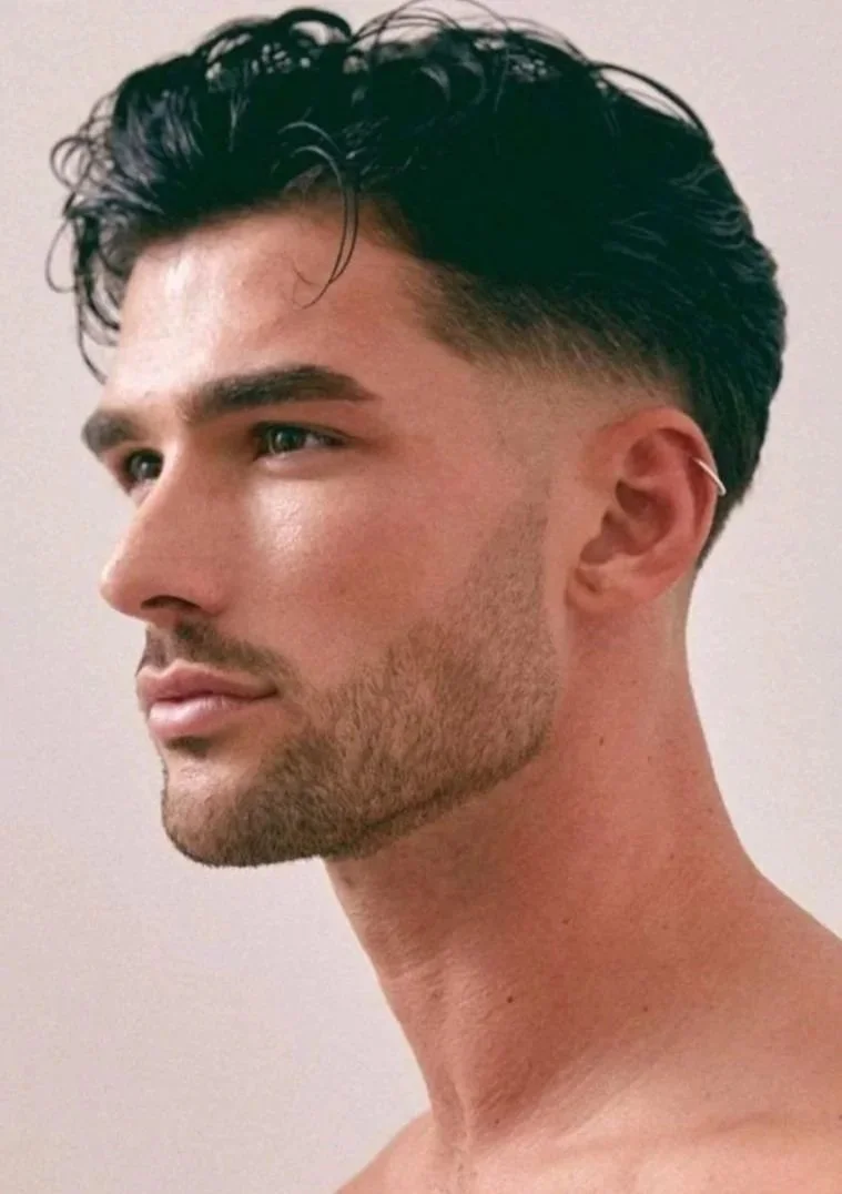 Close-up profile of a young man with dark, styled hair, well-groomed beard, and a small ear cuff, against a plain background.