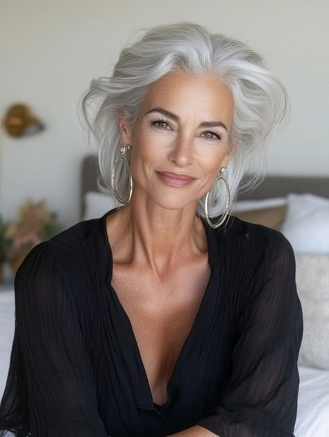 An older woman with silver hair styled in loose waves, wearing large hoop earrings and a black blouse, smiling in a well-lit bedroom.