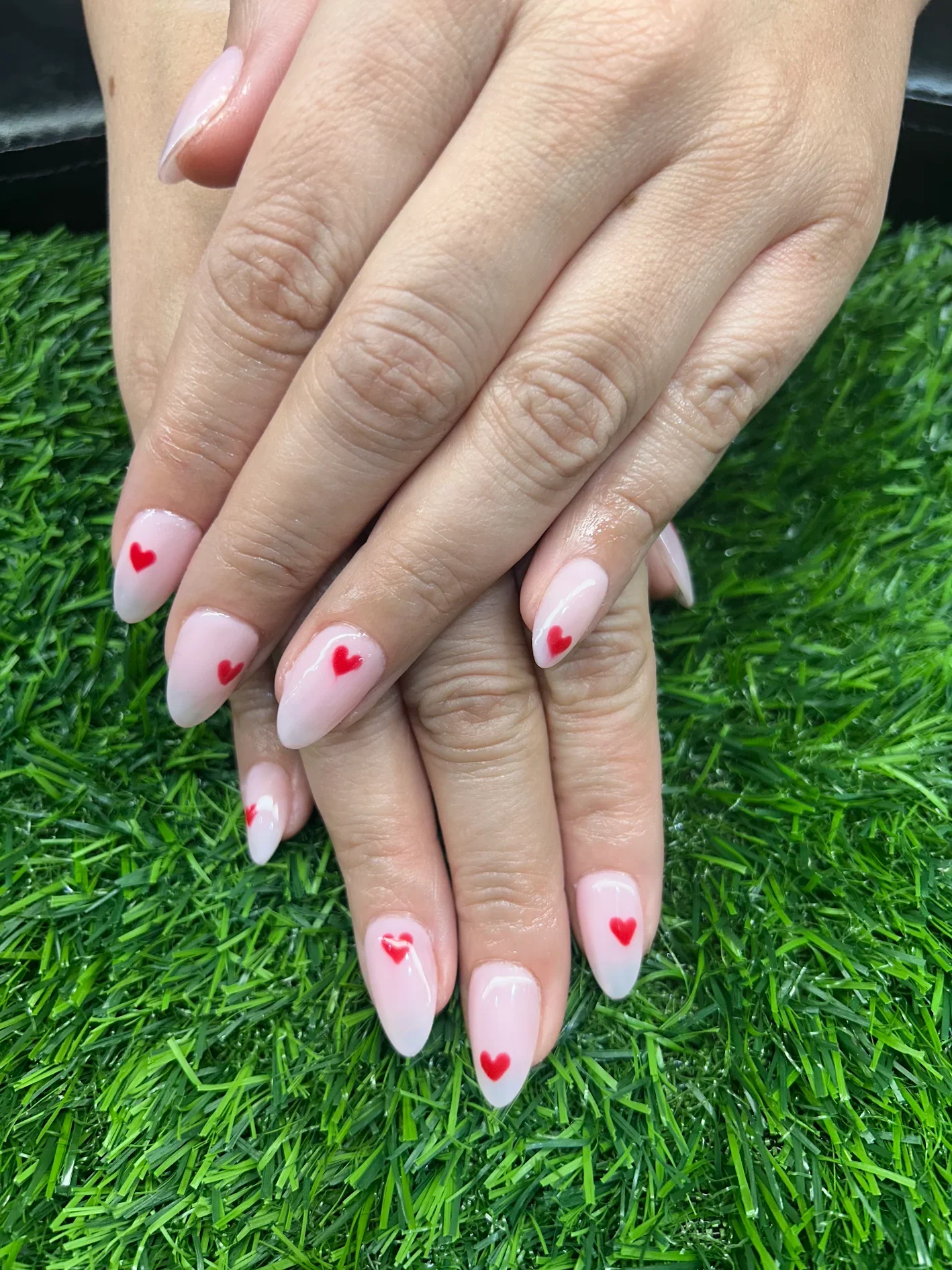 Hands with manicured nails resting on green artificial grass, nails painted with a pale pink base and red heart designs.