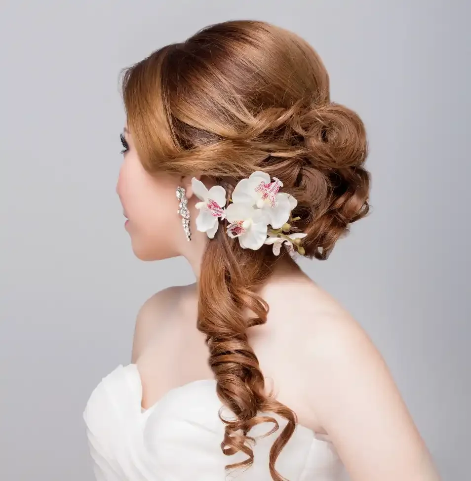 A woman with long, red, curled hair styled in a side-swept manner adorned with white orchids and pearl earrings, wearing a white dress.