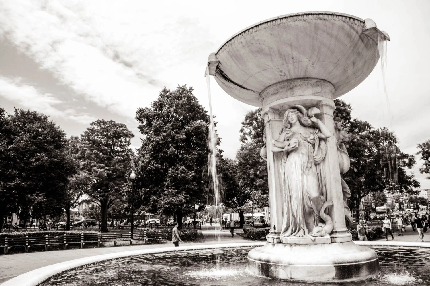 Black and white photograph of a decorative fountain with detailed sculptures of women and animals, situated in a park with trees and people walking in the background.