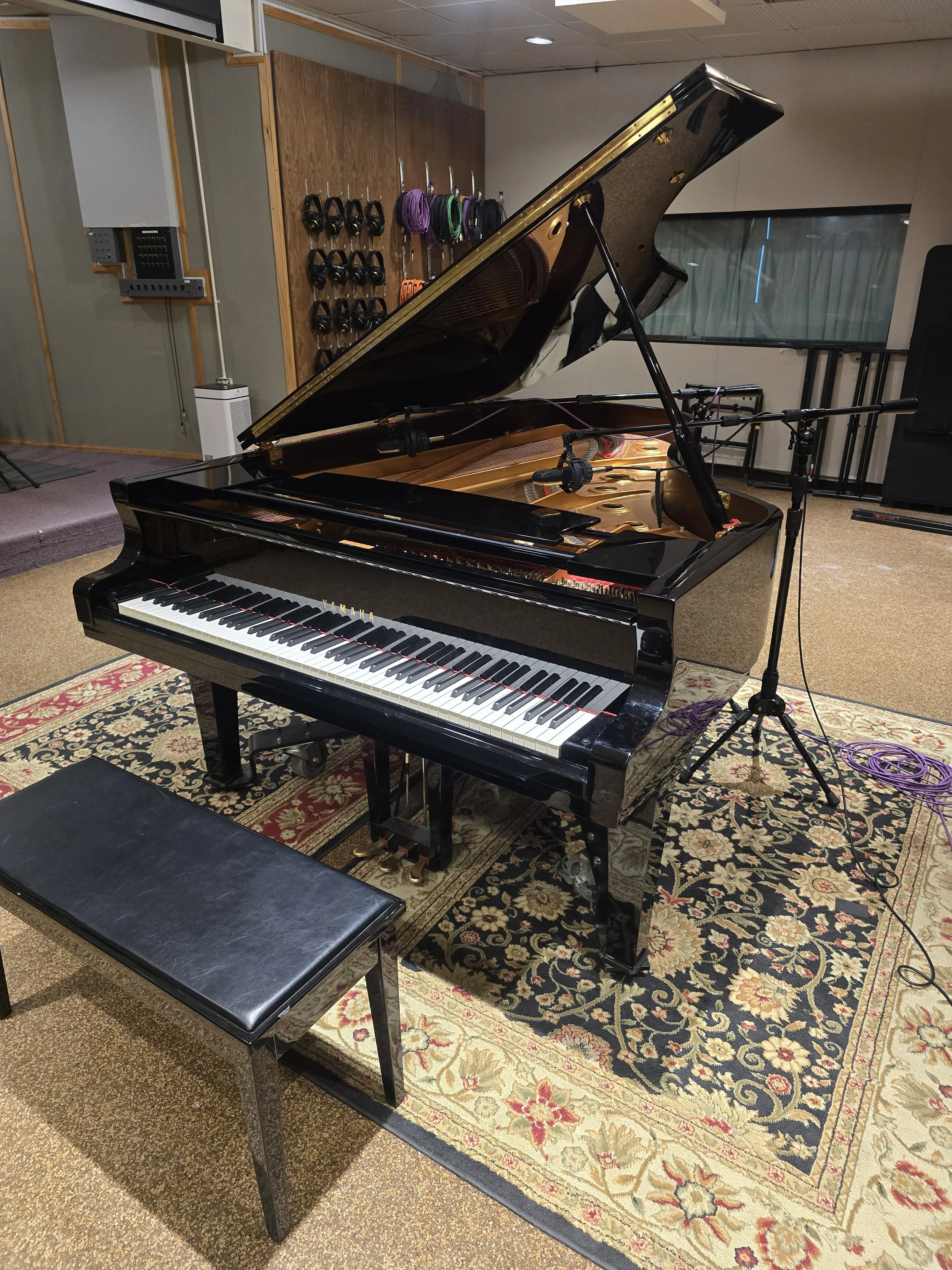 A black grand piano with its lid open, showing the strings and hammers inside, placed on an ornate area rug in a music recording studio. There is a black bench in front of the piano. Behind the piano, on the wall, some soundproofing panels and a rack with headphones and cable organizers are visible.