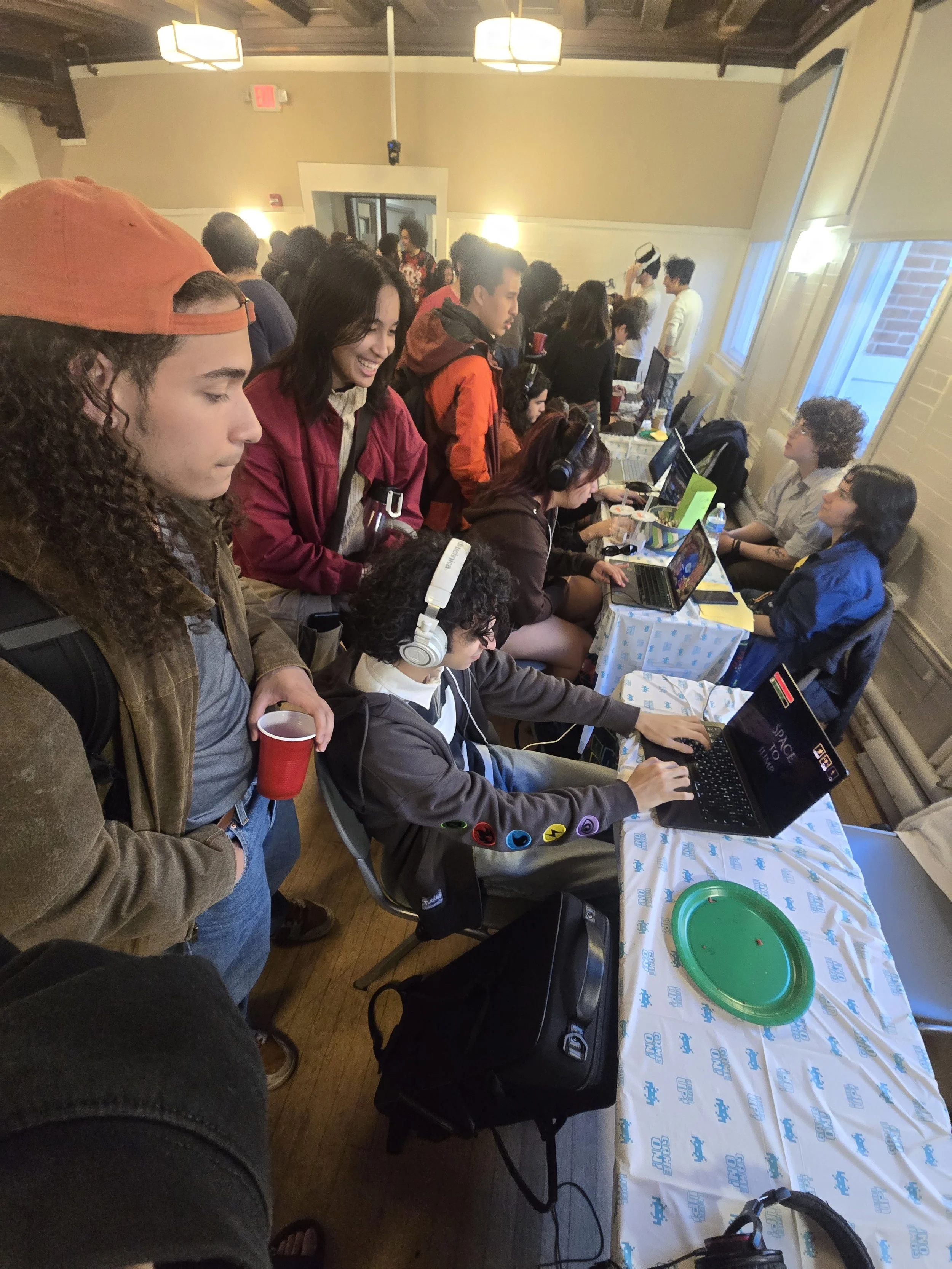 A group of people gathered around a long table, some working on laptops, some talking, with a window and some bright ceiling lights in the background. There are various items like drinks, notebooks, and a green tray on the table.