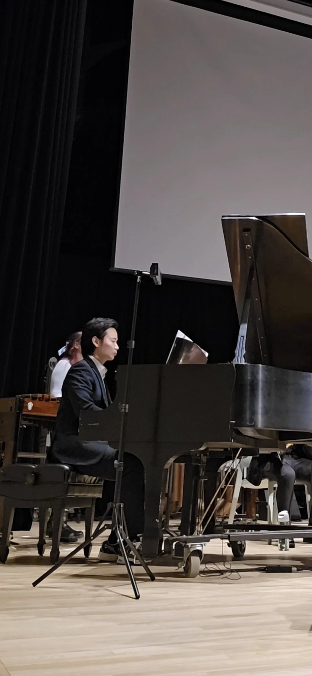 A young man in a black suit playing the piano during a performance in an auditorium.