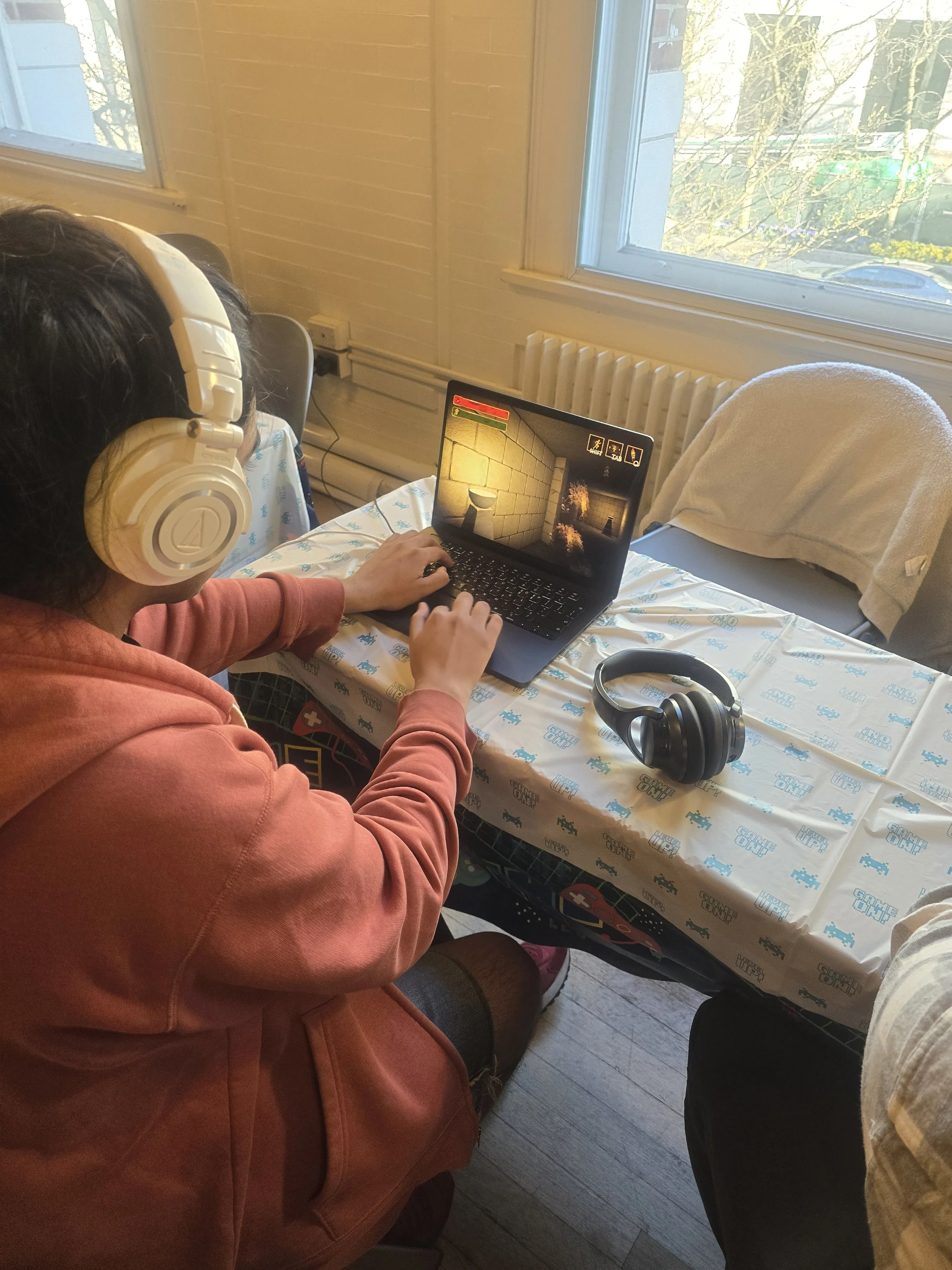 Person wearing a red hoodie and headphones, sitting at a table with a laptop, and working inside a room with large windows and a view of trees outside.