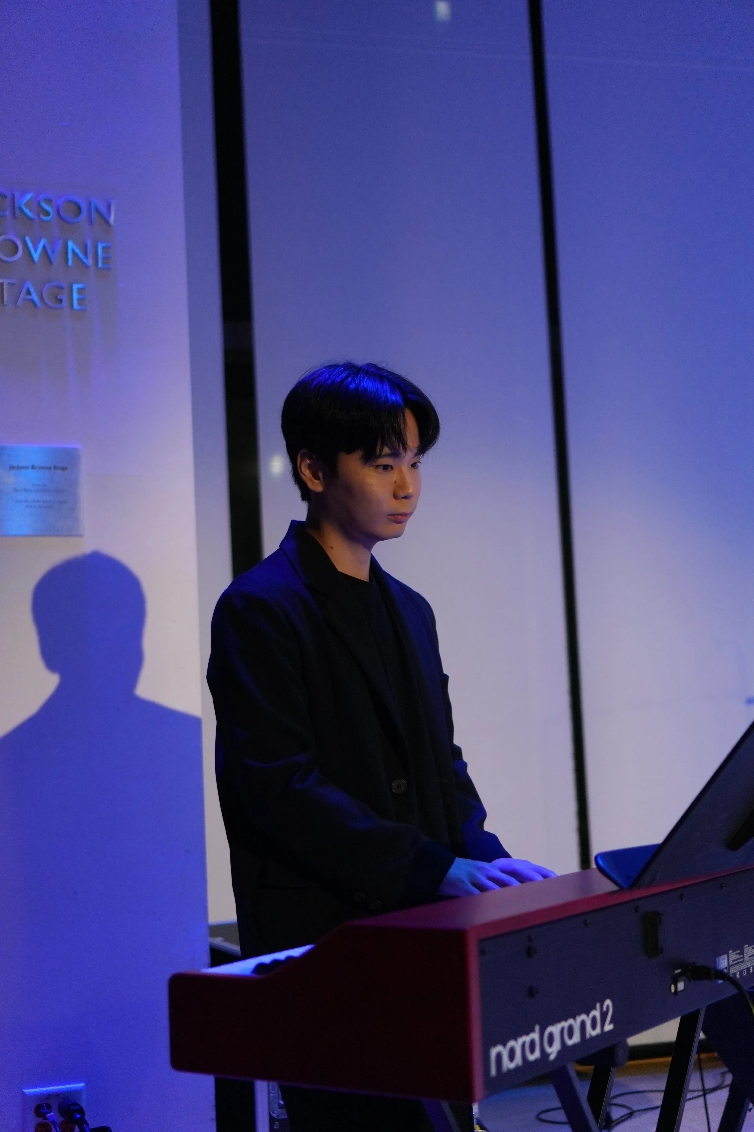 A young man with dark hair playing a red Nord keyboard in a dimly lit room illuminated with blue and purple lighting.