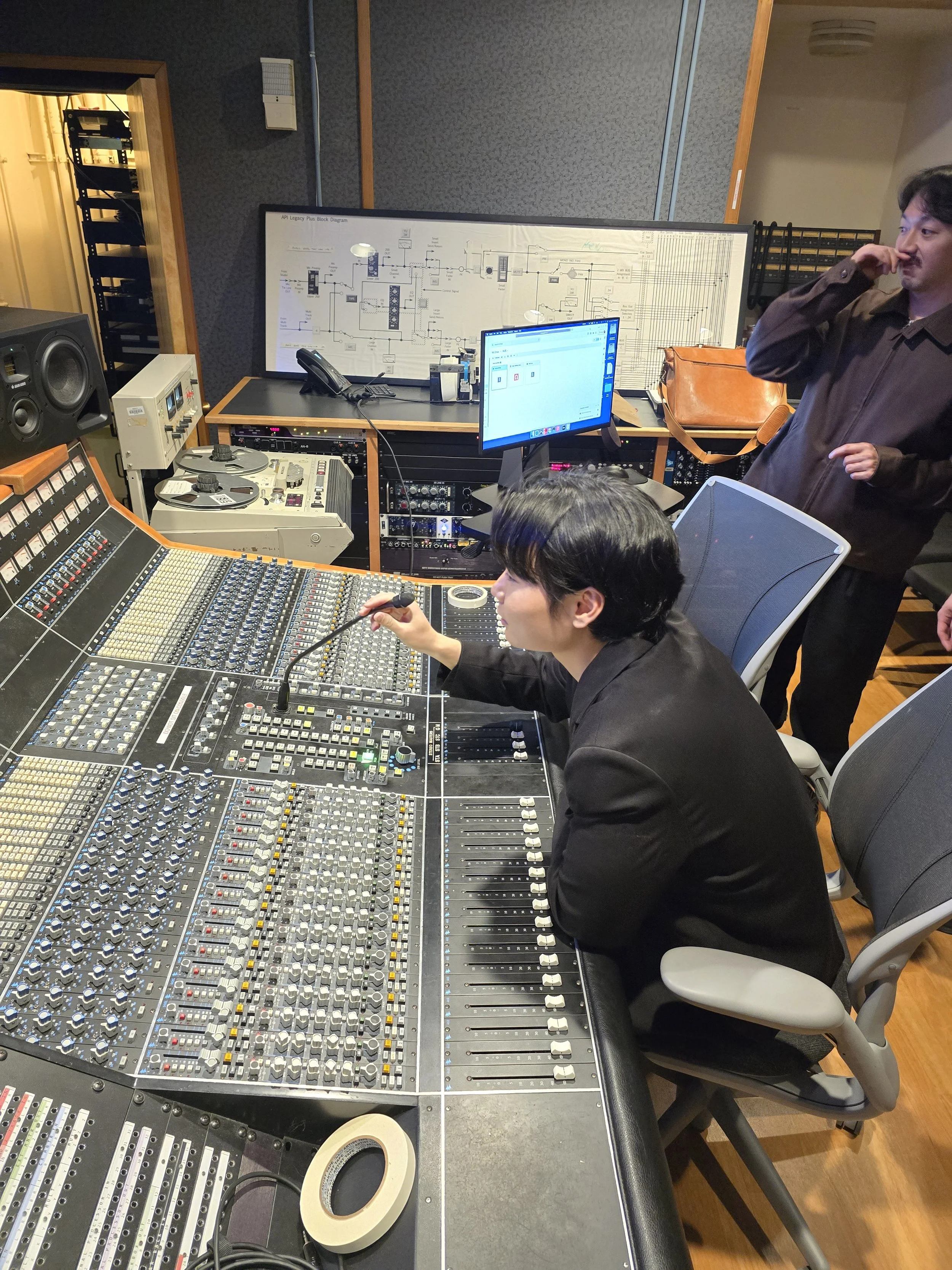 Recording studio with mixing console, two individuals working on technical equipment, and a whiteboard with diagrams.