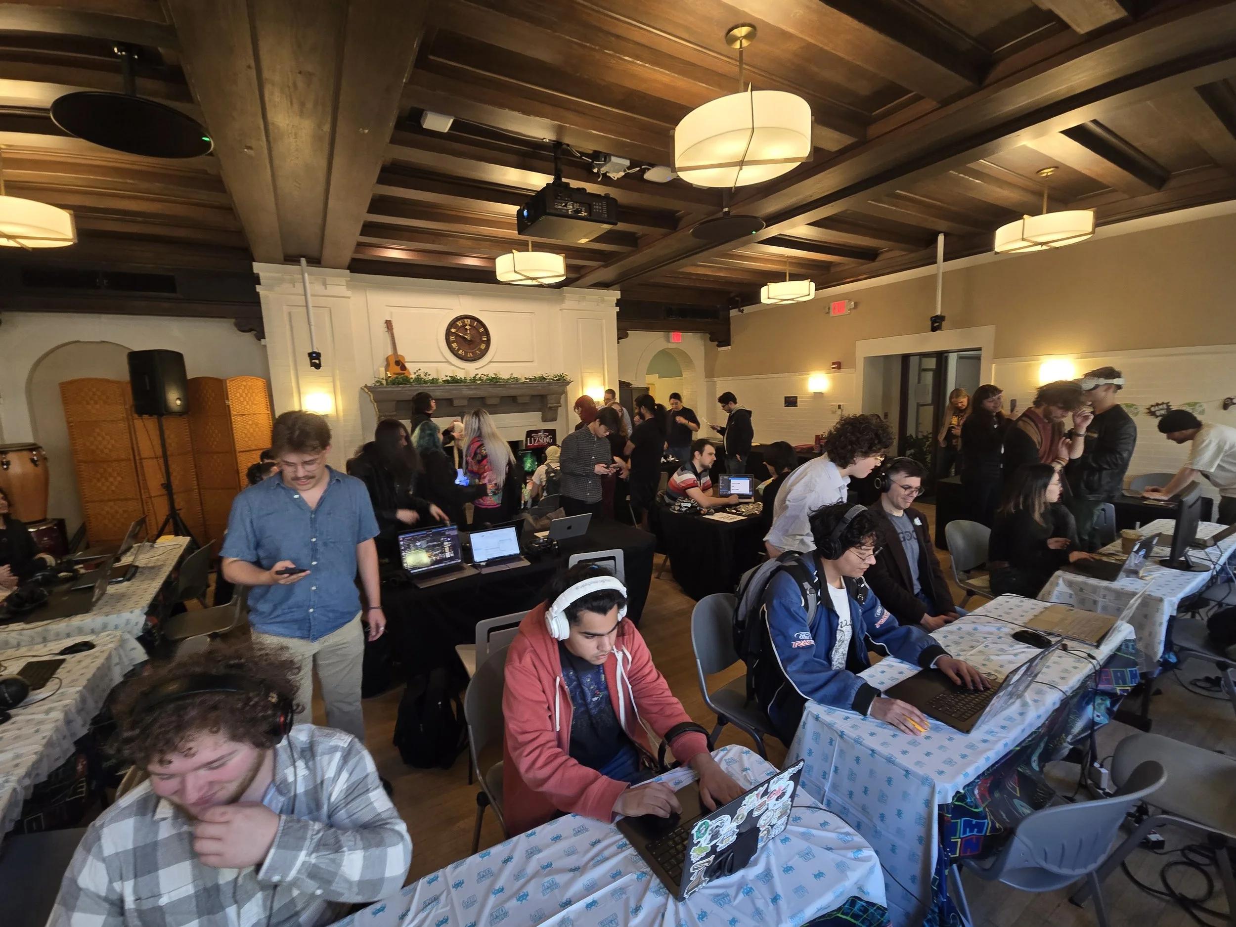 A group of people gathered in a room with a wooden ceiling, some working on laptops or mobile devices, and others standing and talking. The room has a festive atmosphere with tablecloths and decorations, and a clock on the wall above a fireplace.