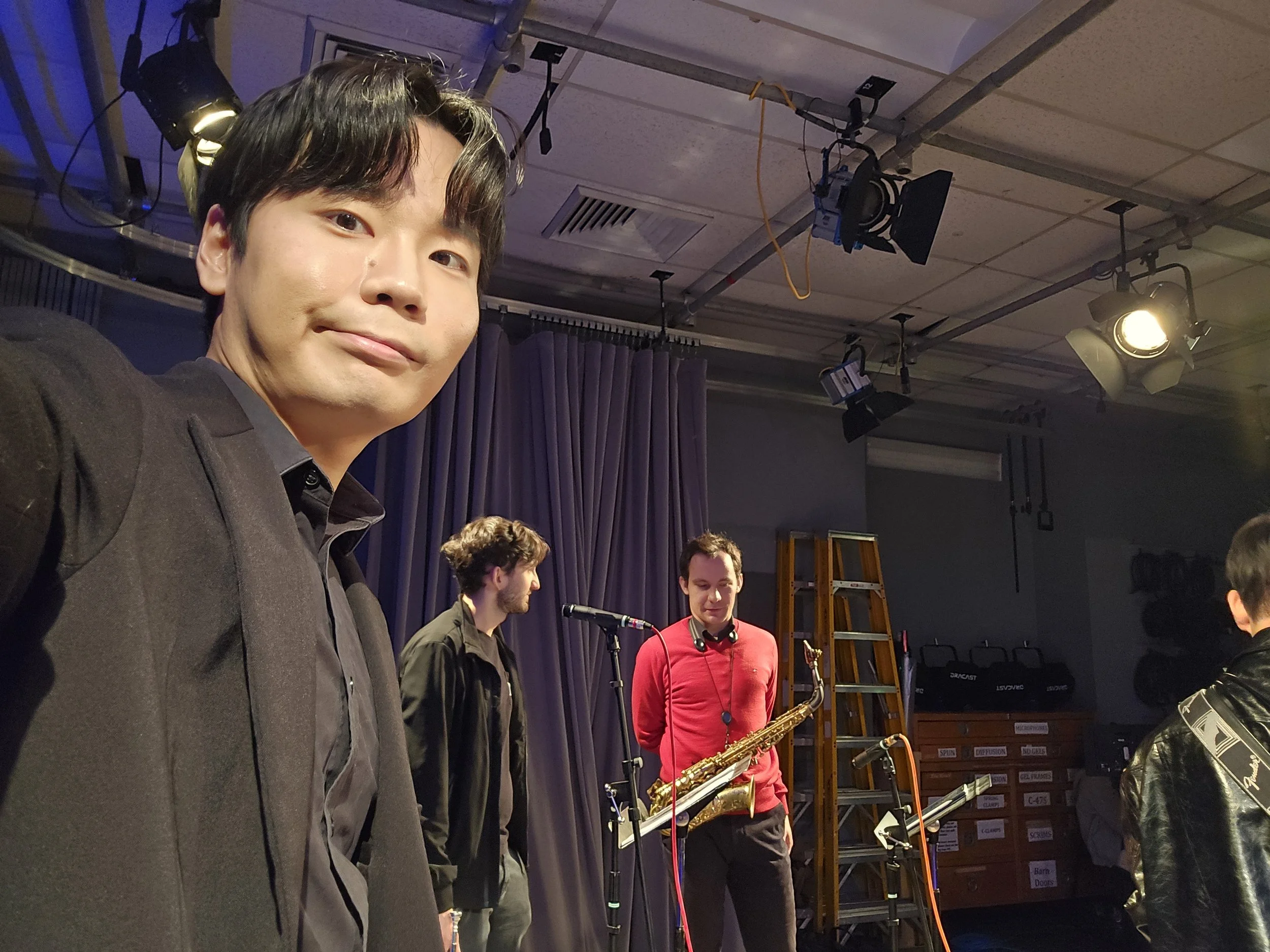 Group of musicians on stage with one person holding a saxophone, backstage or rehearsal setting, studio lighting, and sound equipment visible.