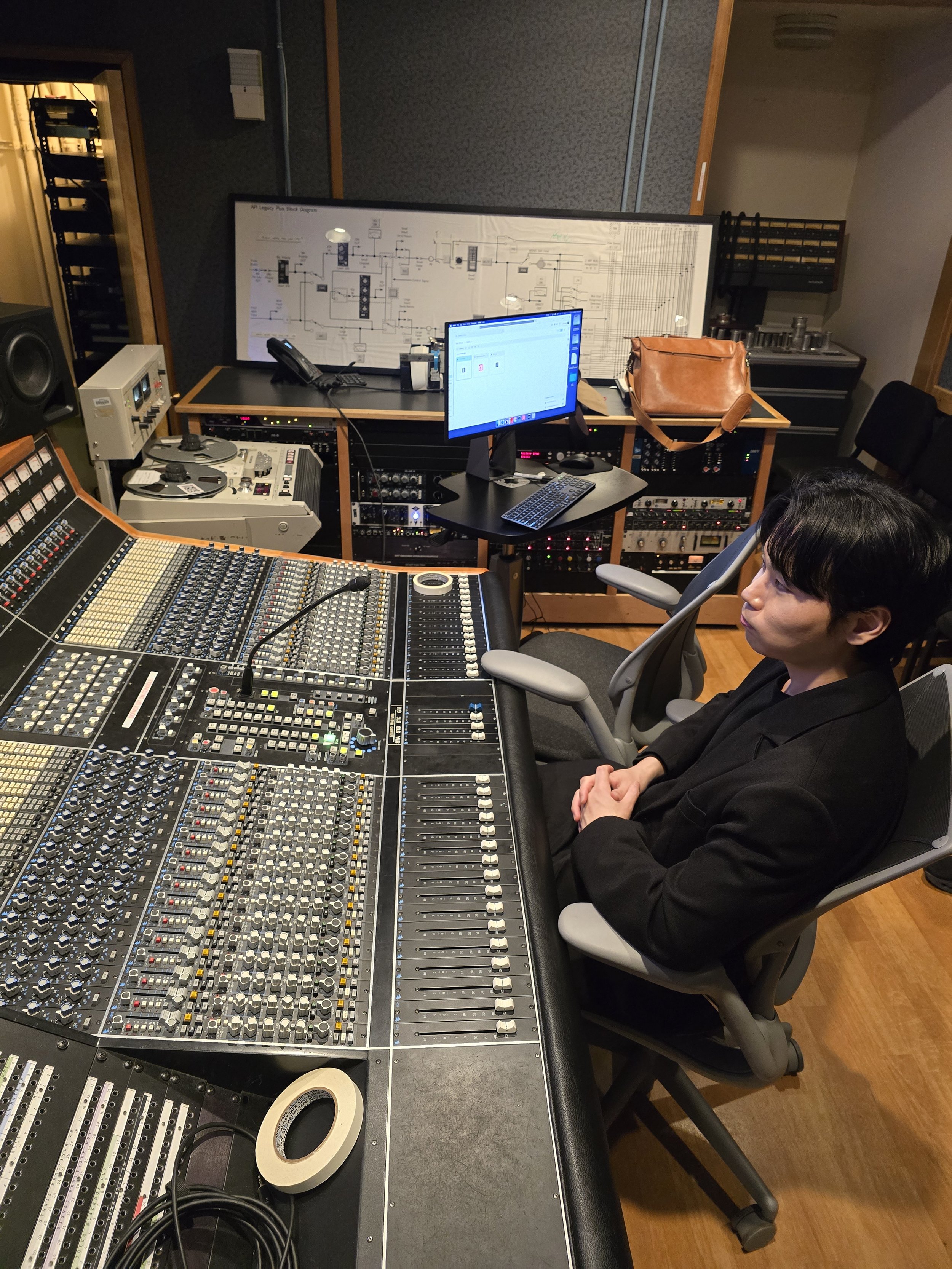 A recording studio with a large audio mixing console, a woman sitting in a chair, and a man sitting in front of the console. There are studio monitors, a computer monitor, and other audio equipment in the room.