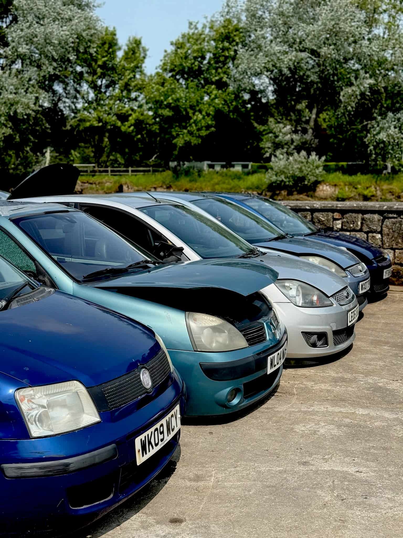 A row of cars parked on a concrete surface with trees and a stone wall in the background.