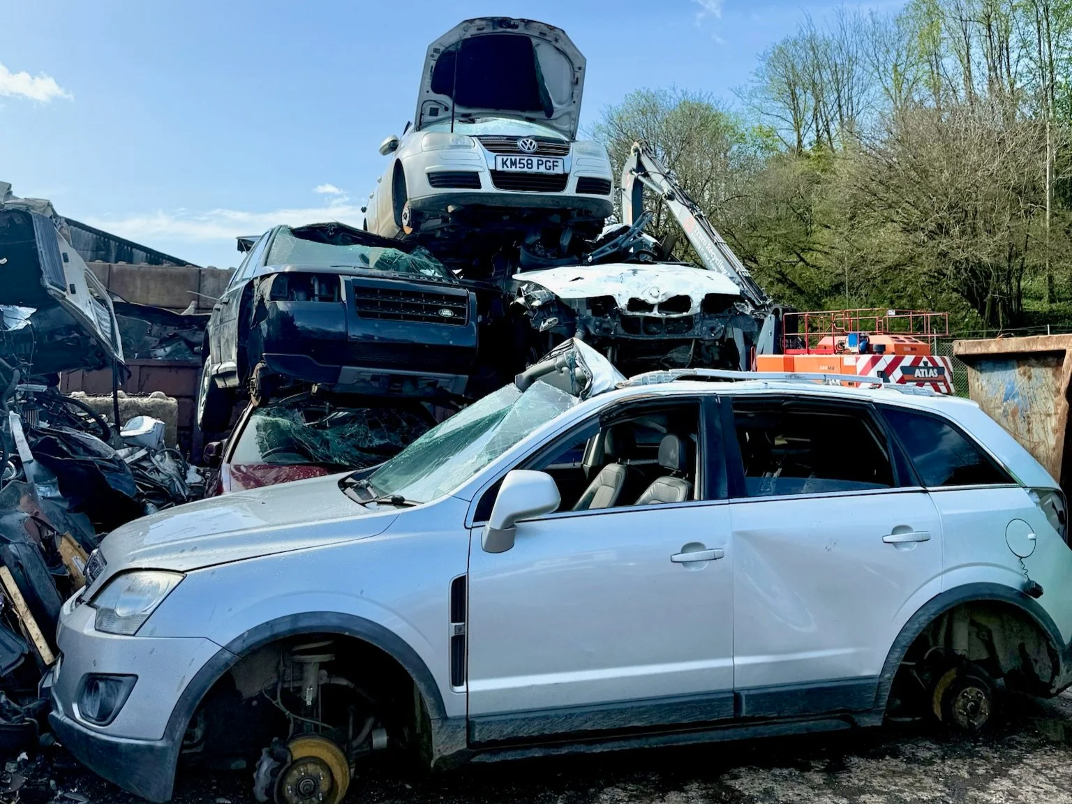 A junkyard with a silver car in the foreground and multiple crushed cars stacked behind it, with a crane visible in the background under a blue sky.