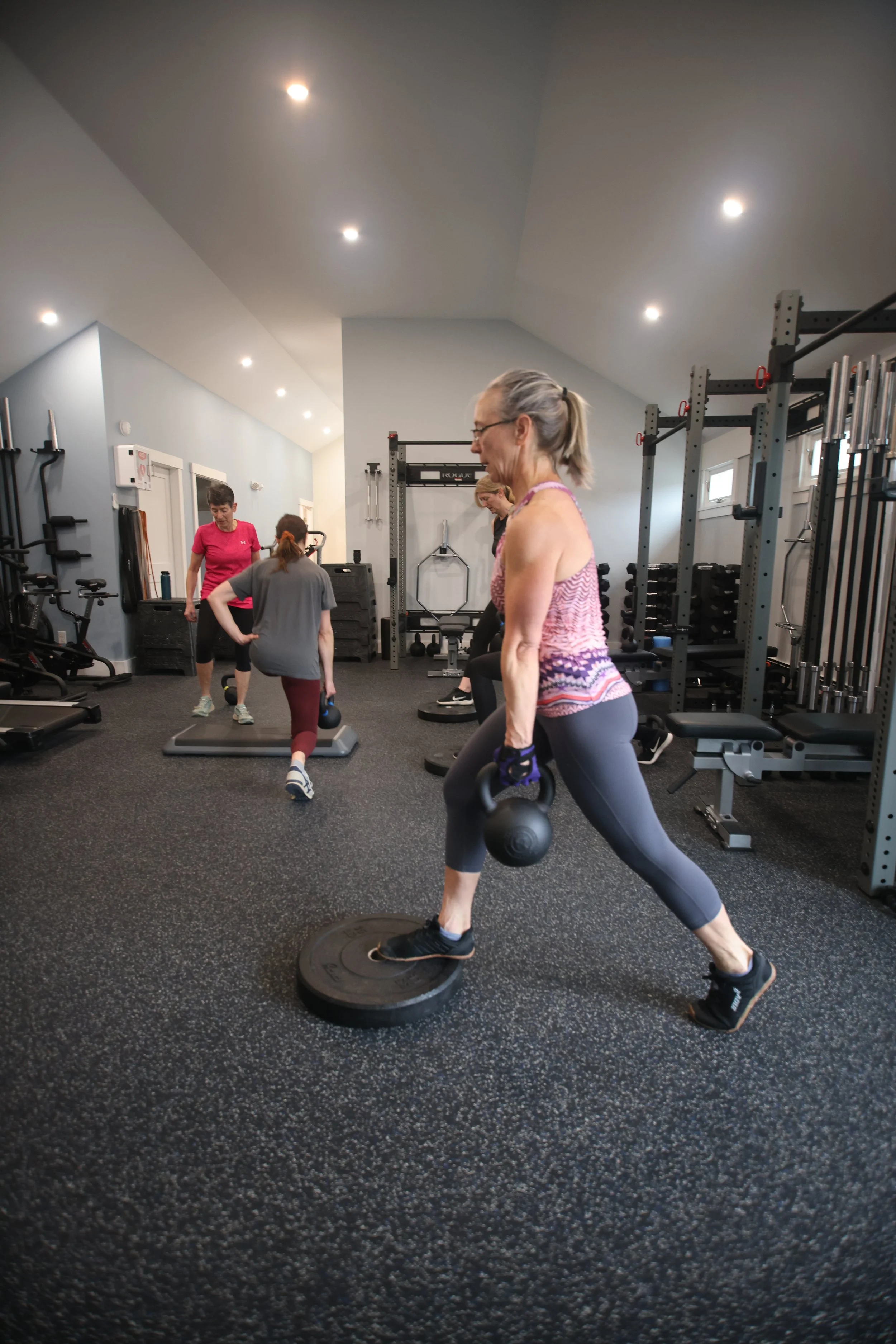 Older woman lifting a kettlebell in a gym with other people exercising in the background.