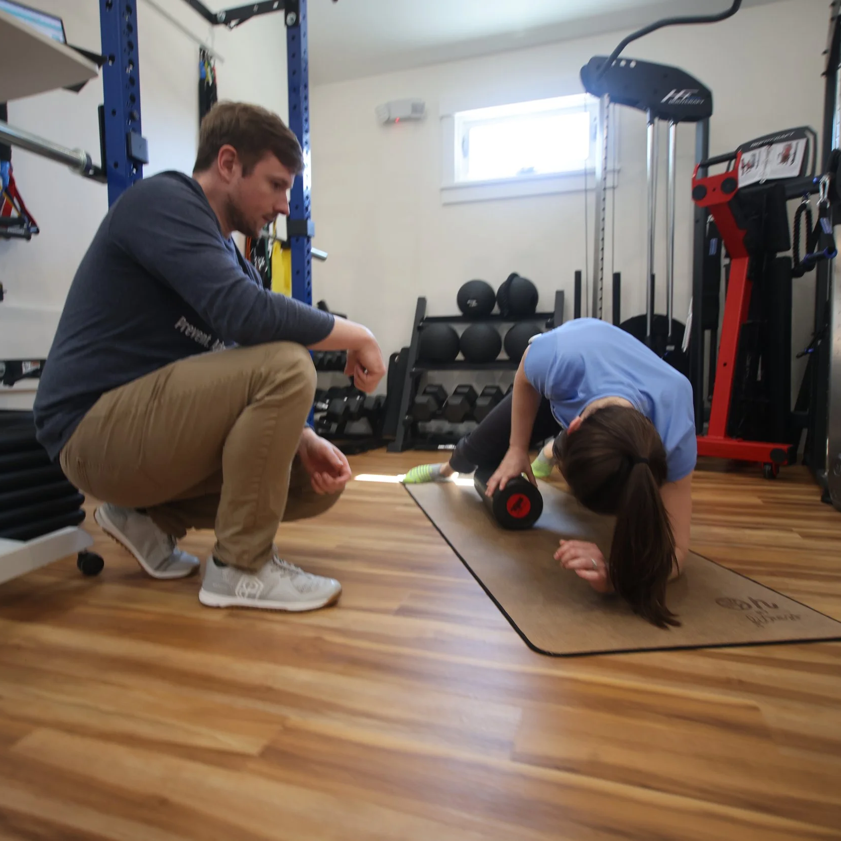 A woman in blue workout clothes is doing an exercise with a foam roller on a yoga mat, while a man in casual clothing observes her in a home gym with various exercise equipment in the background.